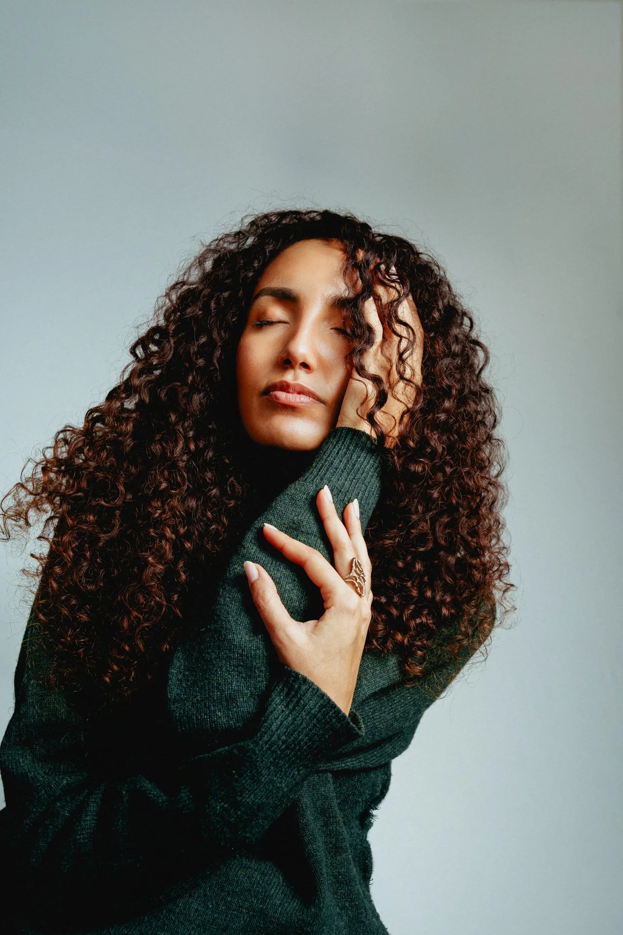 Ragazza con capelli ricci, occhi chiusi, posa contemplativa, indossa maglione verde scuro, copre un orecchio con la mano, sfondo neutro.