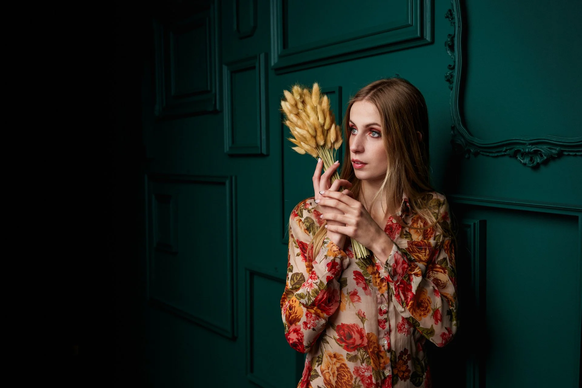 Donna con abito floreale che tiene un mazzo di fiori secchi beige, posa vicino a una parete verde scuro con cornici decorate.