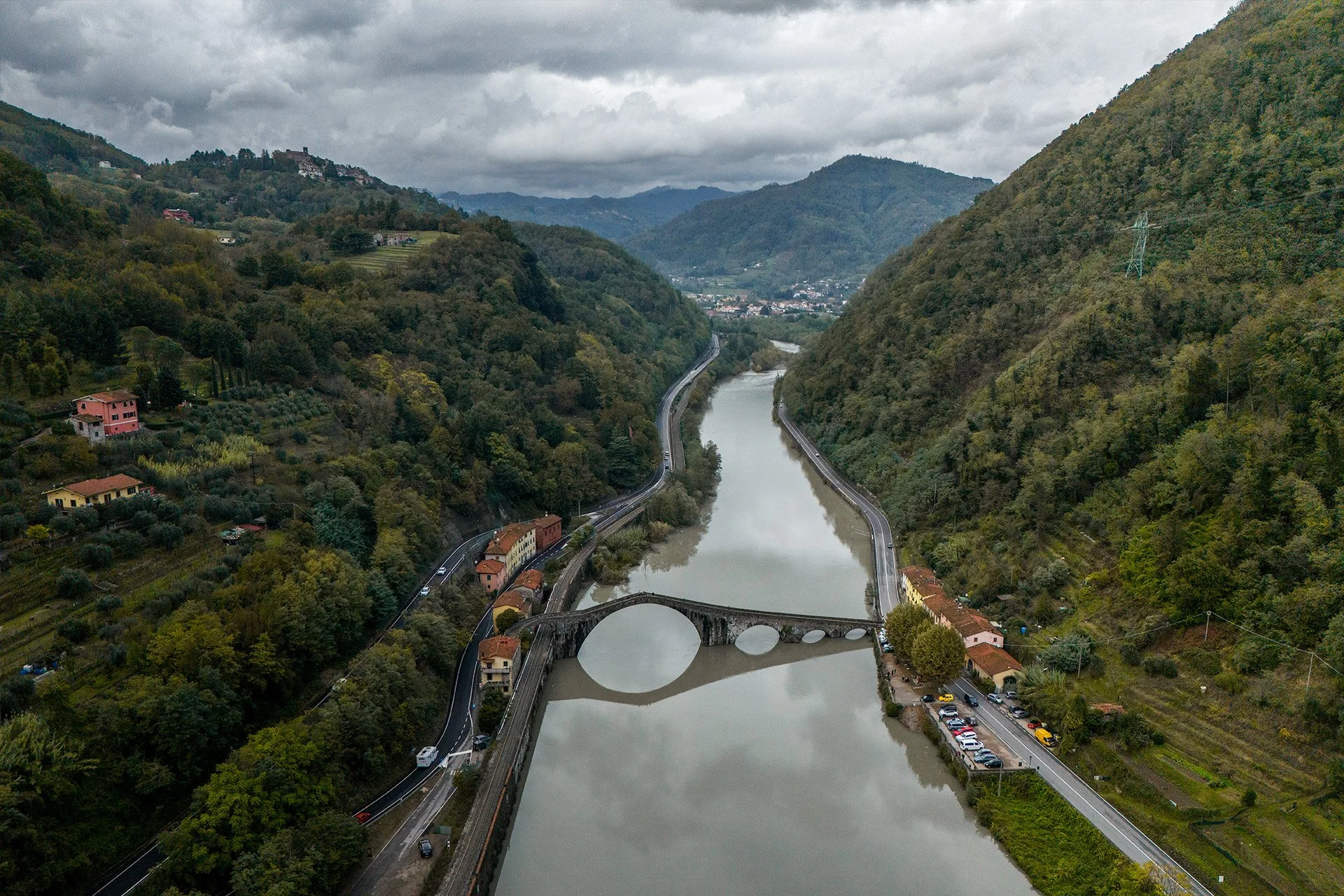 Veduta aerea di un fiume che attraversa una valle tra colline verdi, con un ponte a arcata che attraversa il fiume e strade lungo le rive, case su entrambi i lati e un'area di parcheggio con auto su quella a destra.