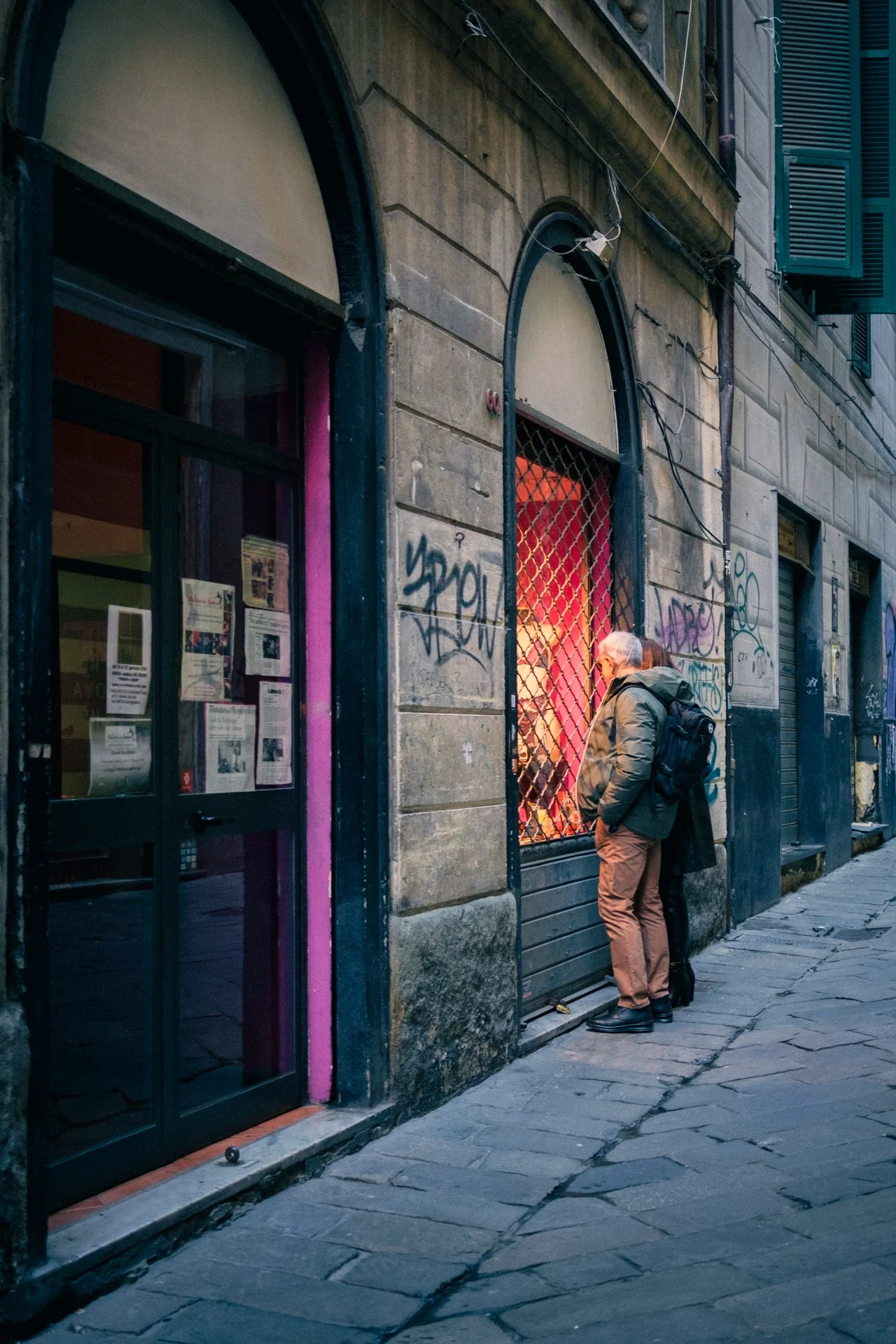 Due persone guardano un negozio con una finestra protetta da una grata metallica, nella strada di una città. La facciata del negozio è decorata con graffiti e i muri sono in pietra, con altri negozi in sottofondo.