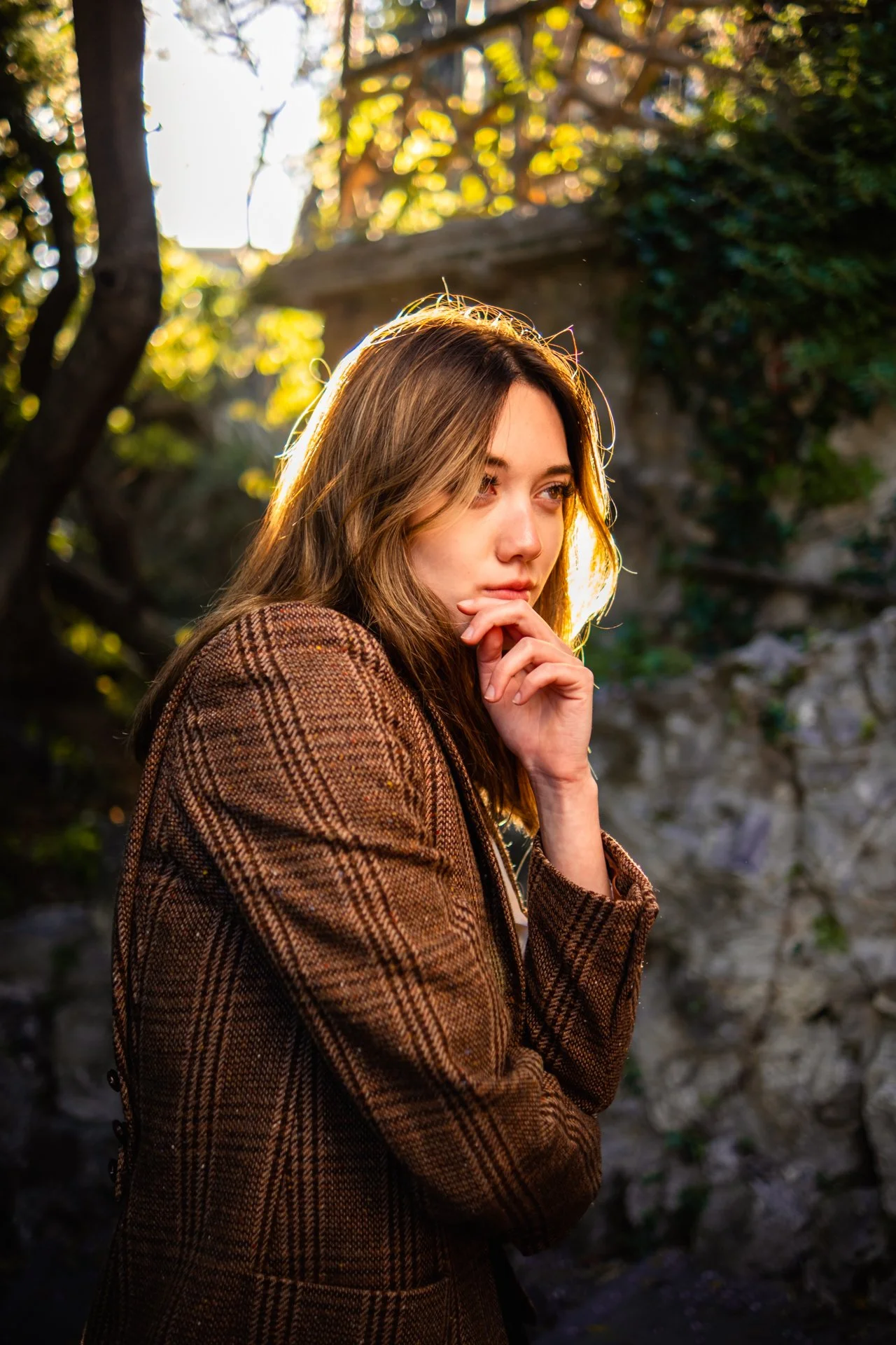 Una donna con capelli castani e vestito a quadri marrone si trova in un ambiente naturale con luci calde e uno sfondo di rocce e vegetazione.