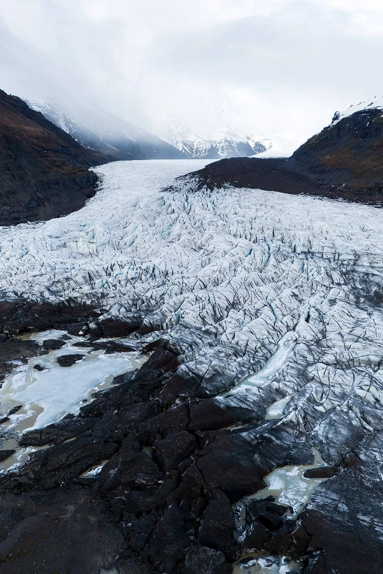 Ghiacciaio tra montagne con neve e rocce scure, cielo nuvoloso.