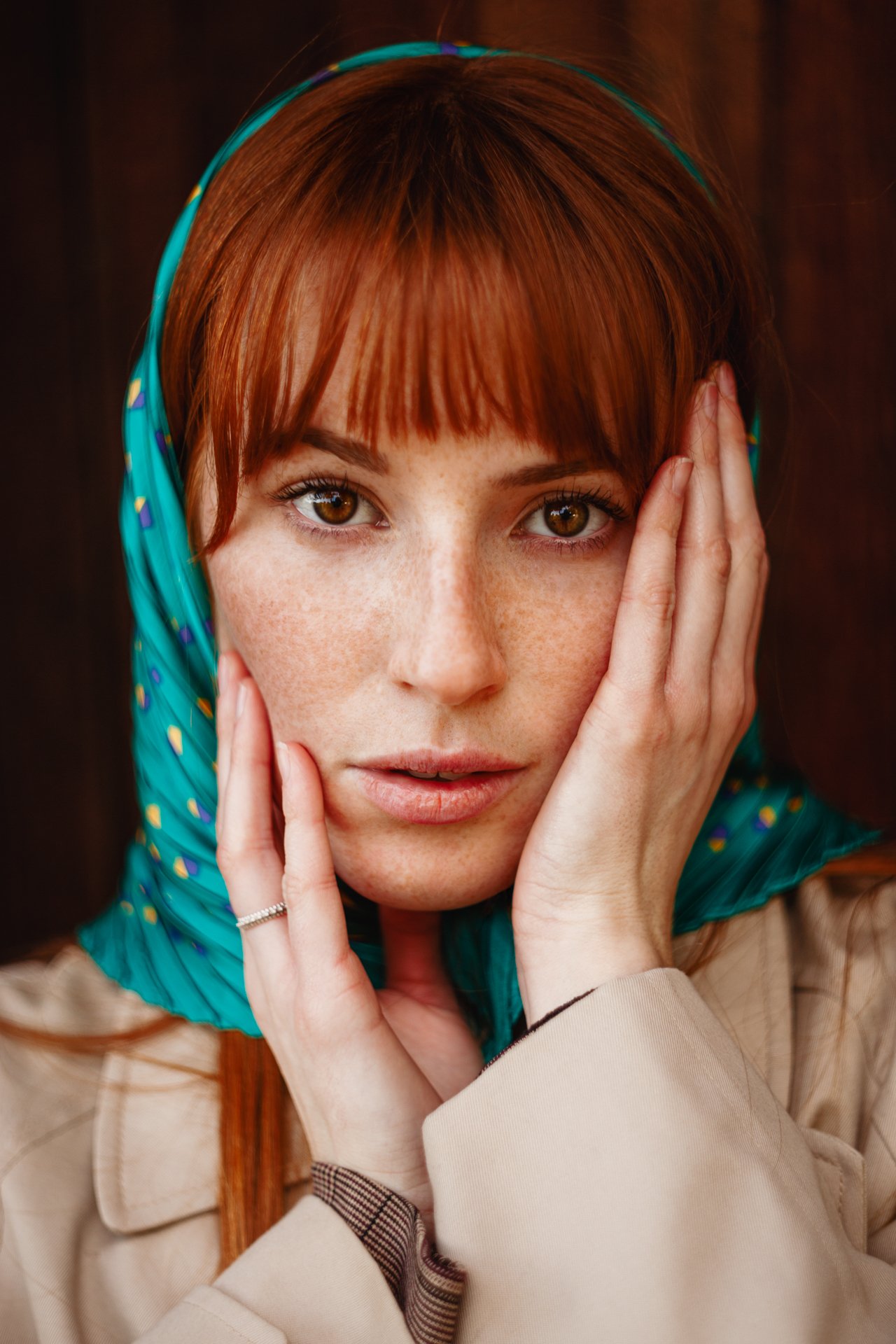 Ragazza con capelli rossi, indossa una sciarpa blu con dettagli colorati e un cappotto beige, con mani che appoggiano sulla faccia, occhi castani, posa in primo piano su sfondo scuro.
