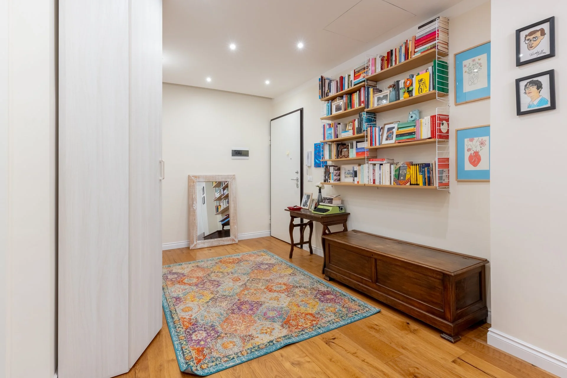 Ingresso di una casa con pareti bianche, libreria con scaffali pieni di libri, tavolo con stampelle verdi, grande specchio con cornice chiara, armadio bianco, tappeto colorato e riffa di quadrini e disegni alle pareti.