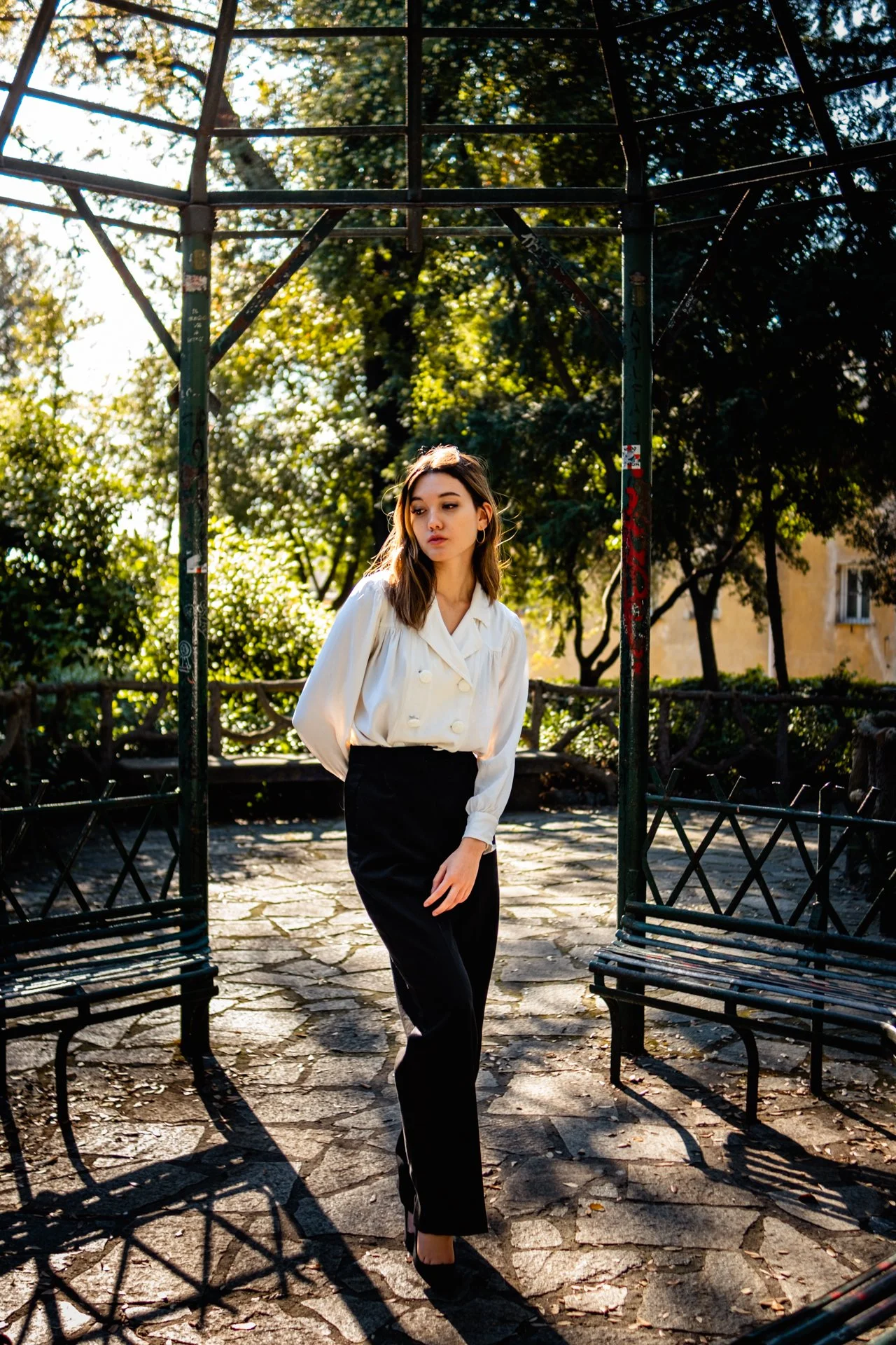 Una giovane donna con capelli castani, vestita con una camicetta bianca e pantaloni neri, si trova in un parco soleggiato tra panchine di metallo, circondata da alberi e vegetazione.