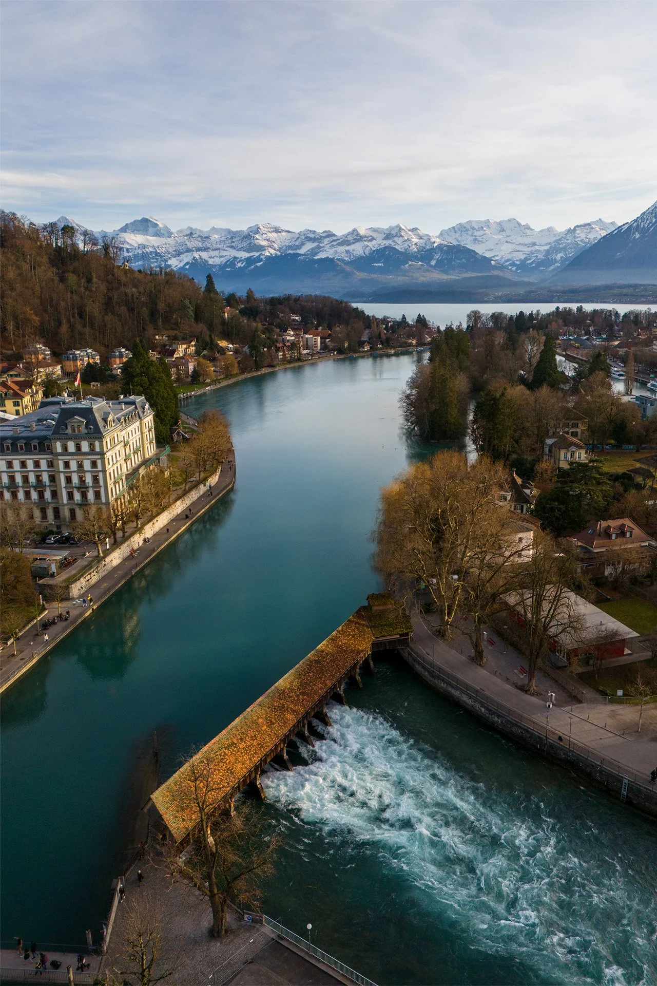 Vista aerea di un lago circondato da montagne innevate e un paesaggio urbano, con edifici, alberi e un fiume che scorre attraverso la città.