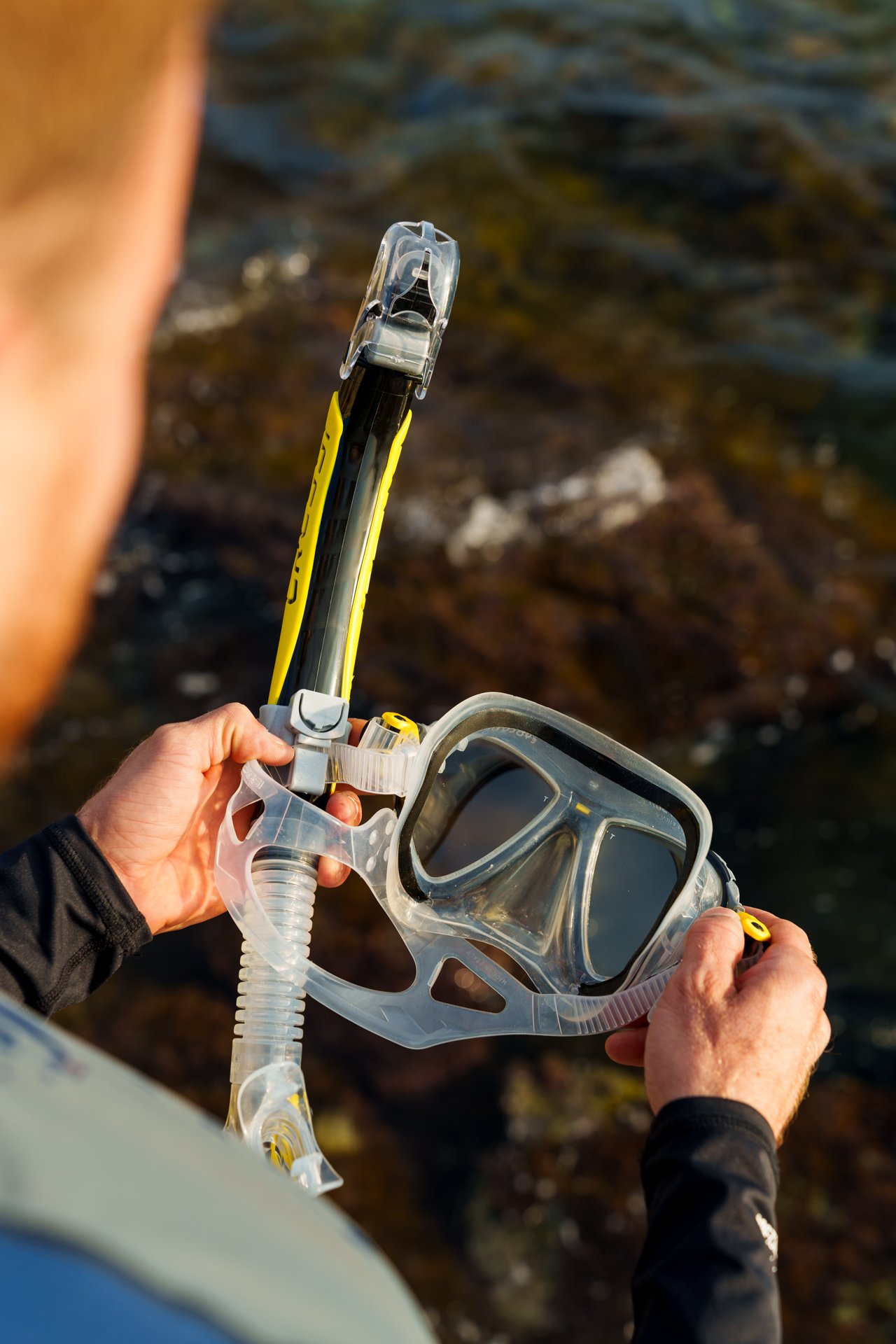 Persona che tiene in mano una maschera da sub con un paio di pinne gialle in un ambiente di spiaggia o vicino all'acqua.