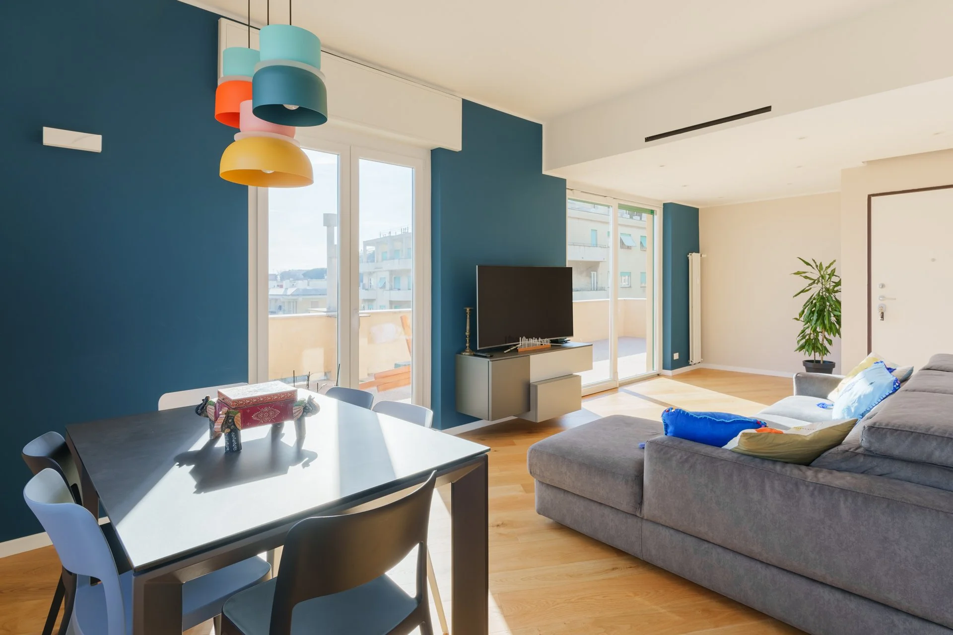 Soggiorno con parete blu, divano grigio, tavolo da pranzo e televisore, grandi finestre che danno sul balcone e illuminano la stanza.