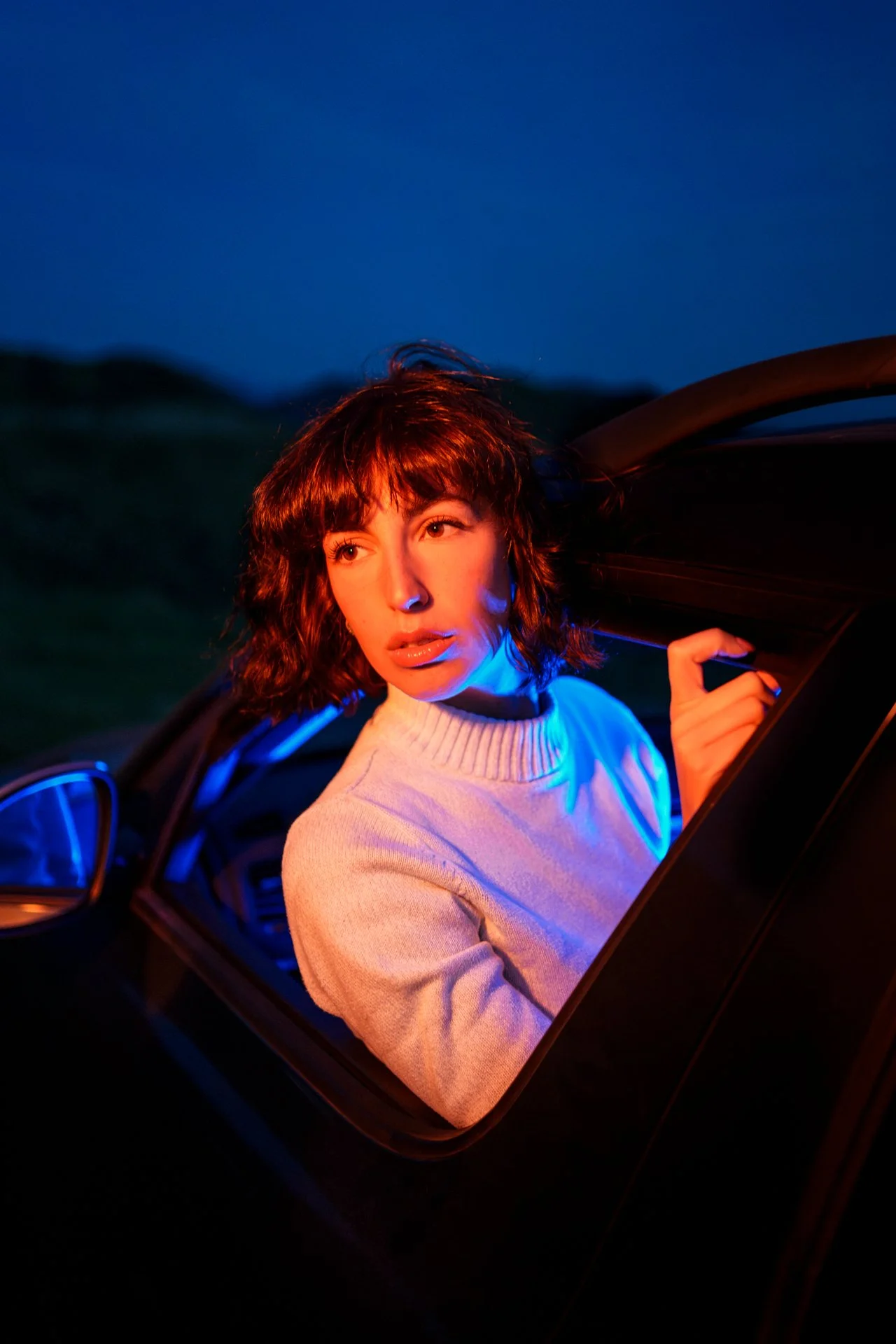 Una donna con capelli ricci e vestito chiaro che si affaccia dal finestrino di un'auto al tramonto o di notte, illuminata da luci colorate.