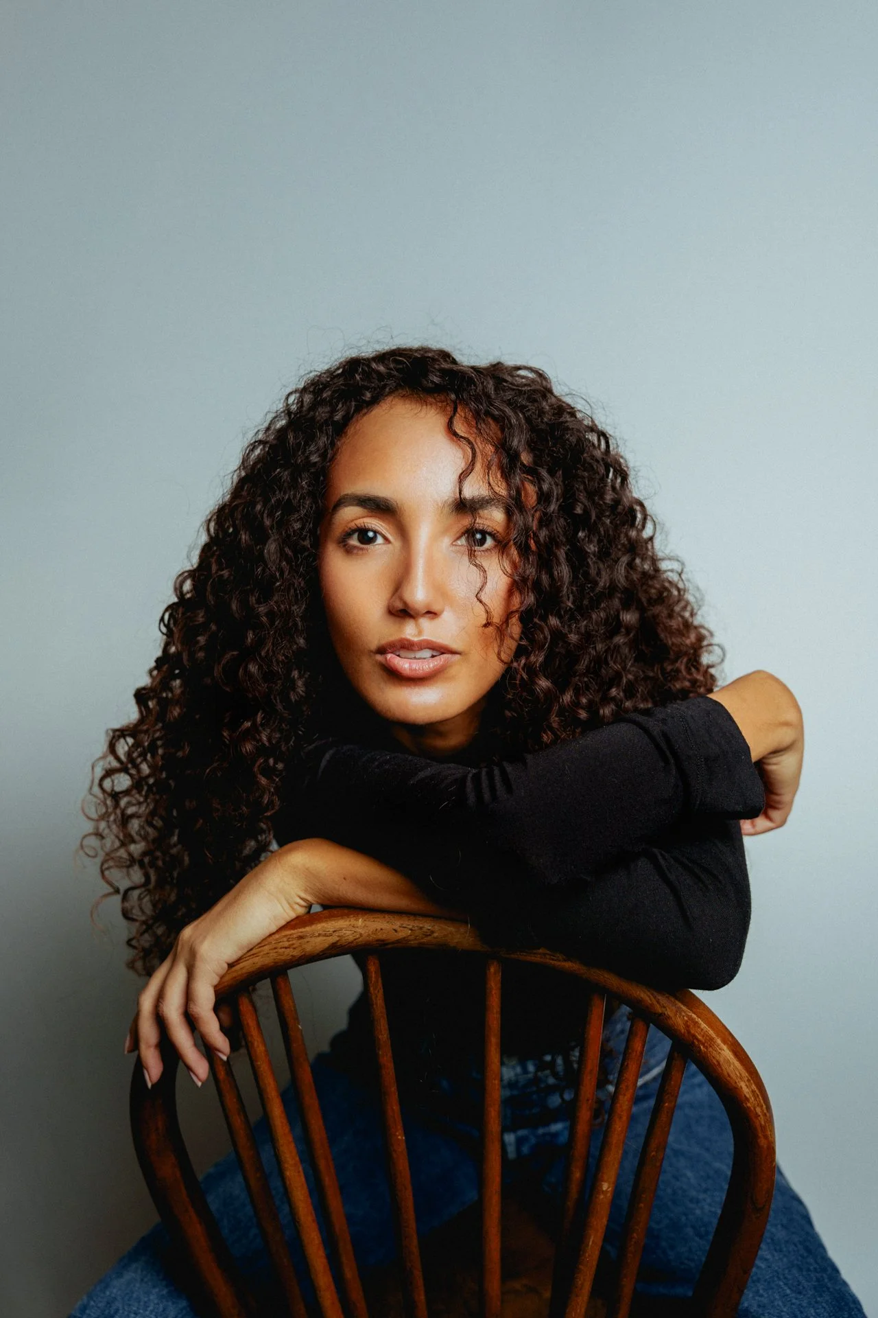 Ragazza con capelli ricci seduta su una sedia di legno, con braccia appoggiate sulla schiena della sedia, indossa una maglietta nera e jeans.