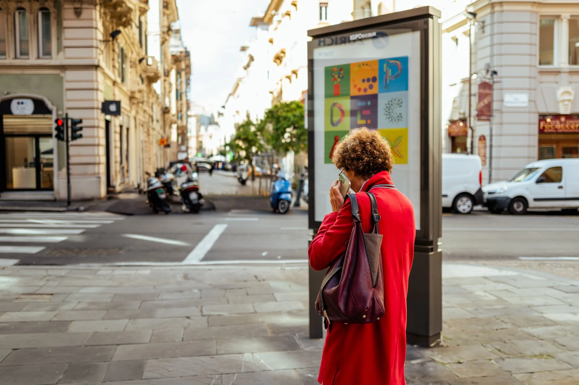 Donna anziana con cappotto rosso e borsa in spalla parla al telefono sulla strada in una città