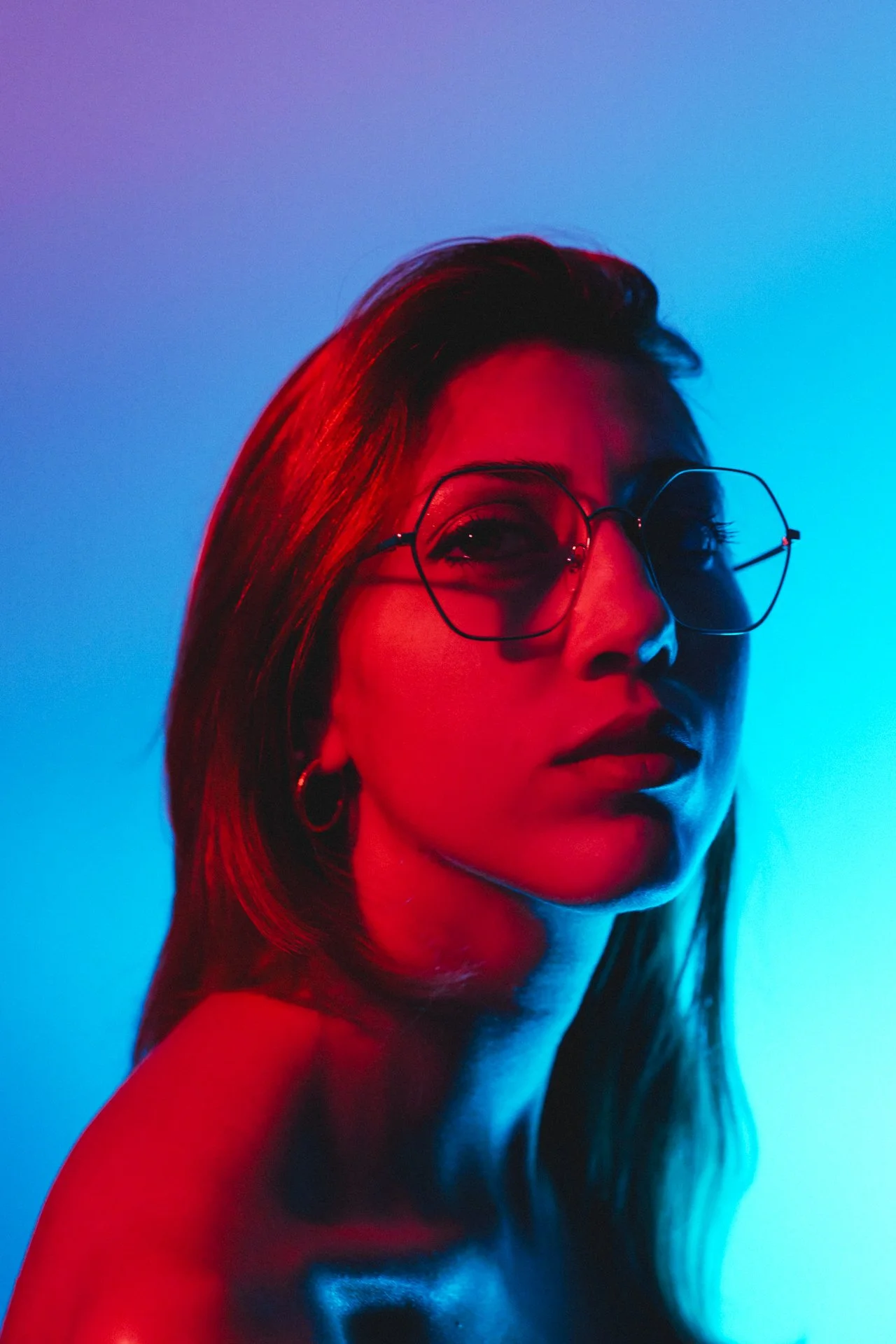 Ritratto di una donna con capelli lunghi, occhiali da vista a forma di esagono e orecchini, illuminata con luci rosse e blu