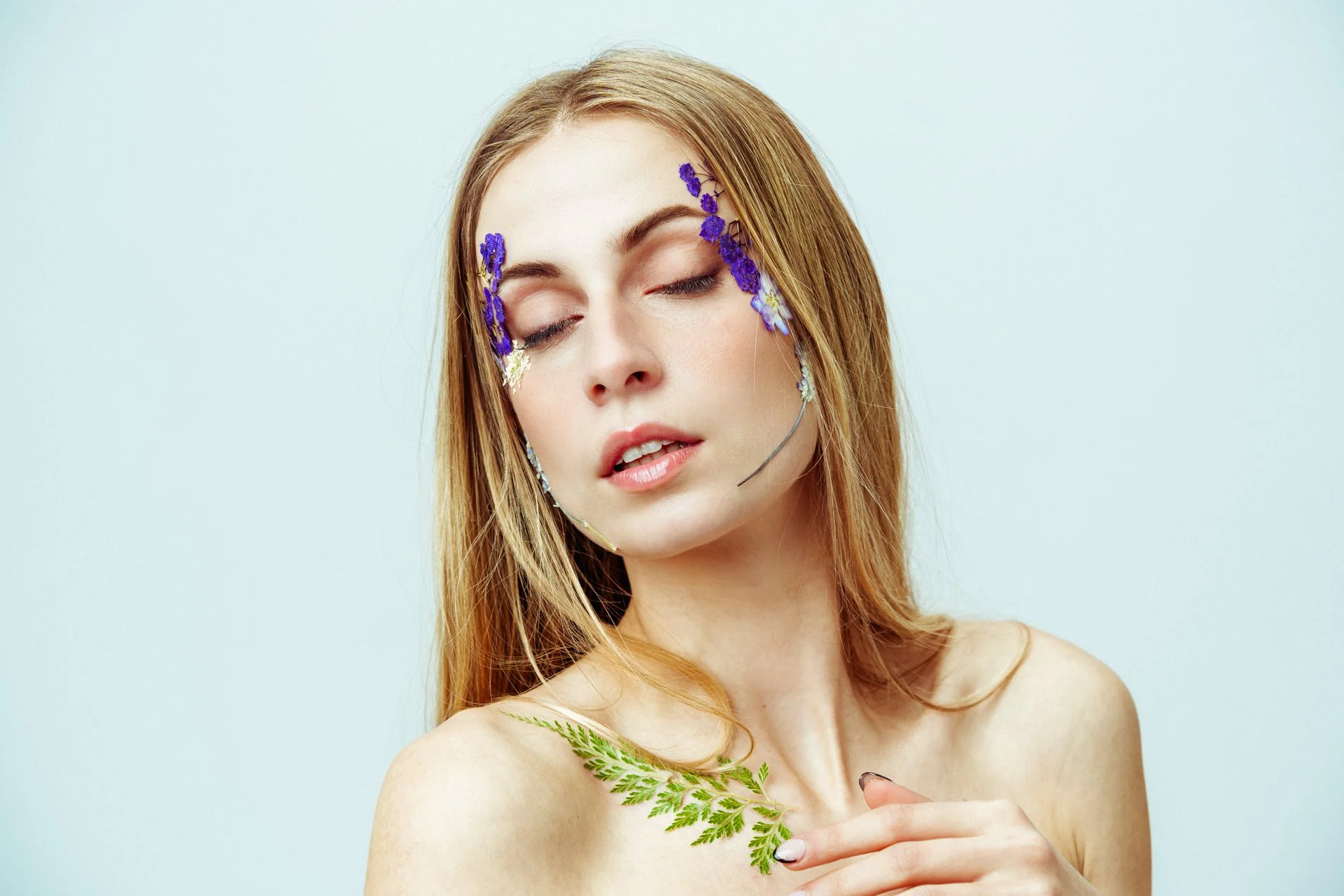 Ragazza con capelli rossi e occhi chiusi, decorata con fiori viola e bianchi sul viso, che tiene una foglia di felce sulla spalla, sfondo chiaro.