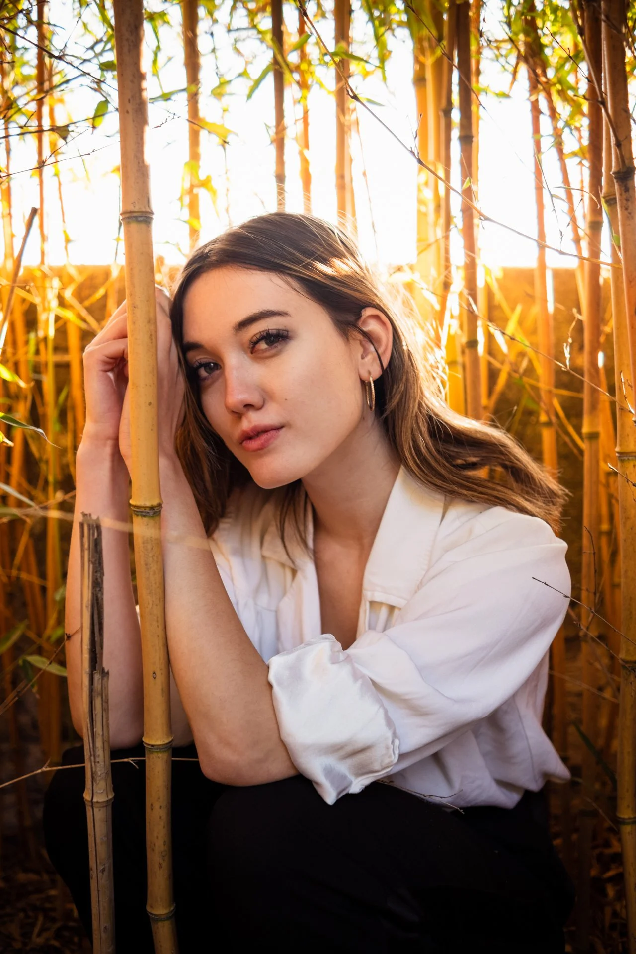 Ragazza con capelli castani in una posizione seduta tra canne di bambù, illuminata dalla luce dorata del tramonto.