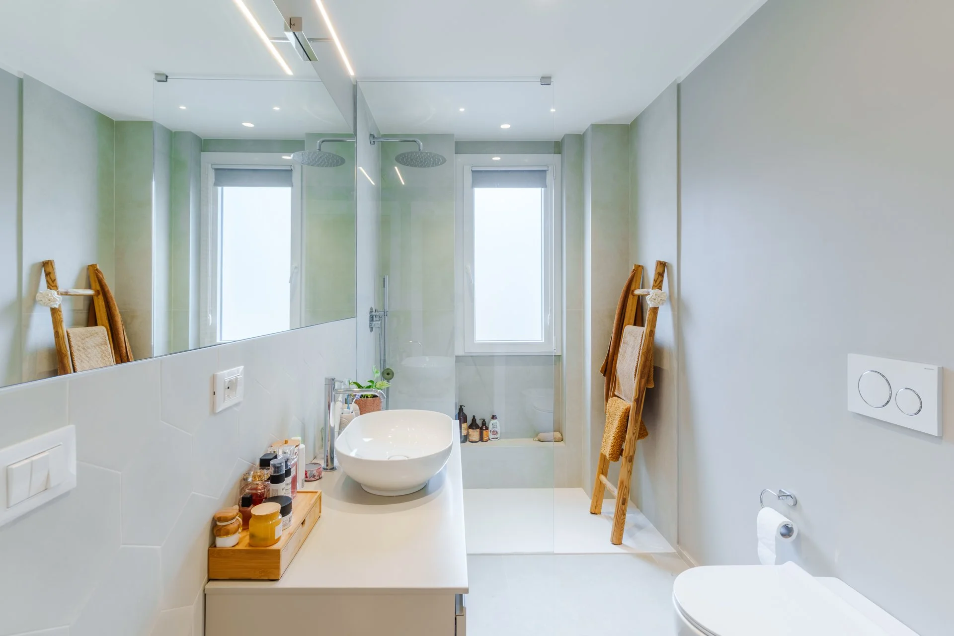Bagno modernamente arredato con lavabo, specchio grande, doccia con trasparente pareti di vetro, luci a soffitto, finestra e asciugamani di tessuto appesi su una sedia in legno.