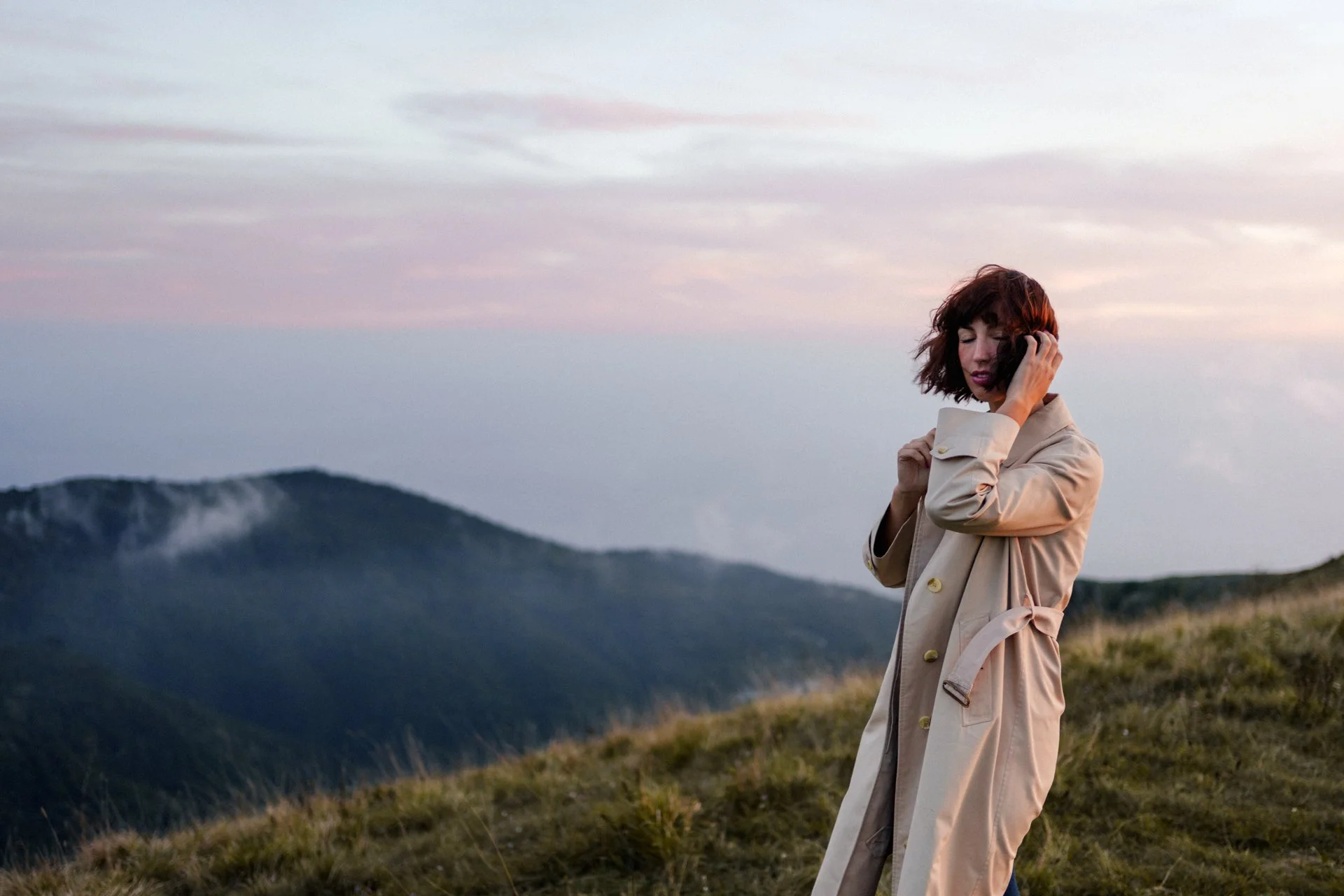Donna con cappotto beige si trova su una collina durante il tramonto, con montagne sullo sfondo e nuvole rosa e viola nel cielo.