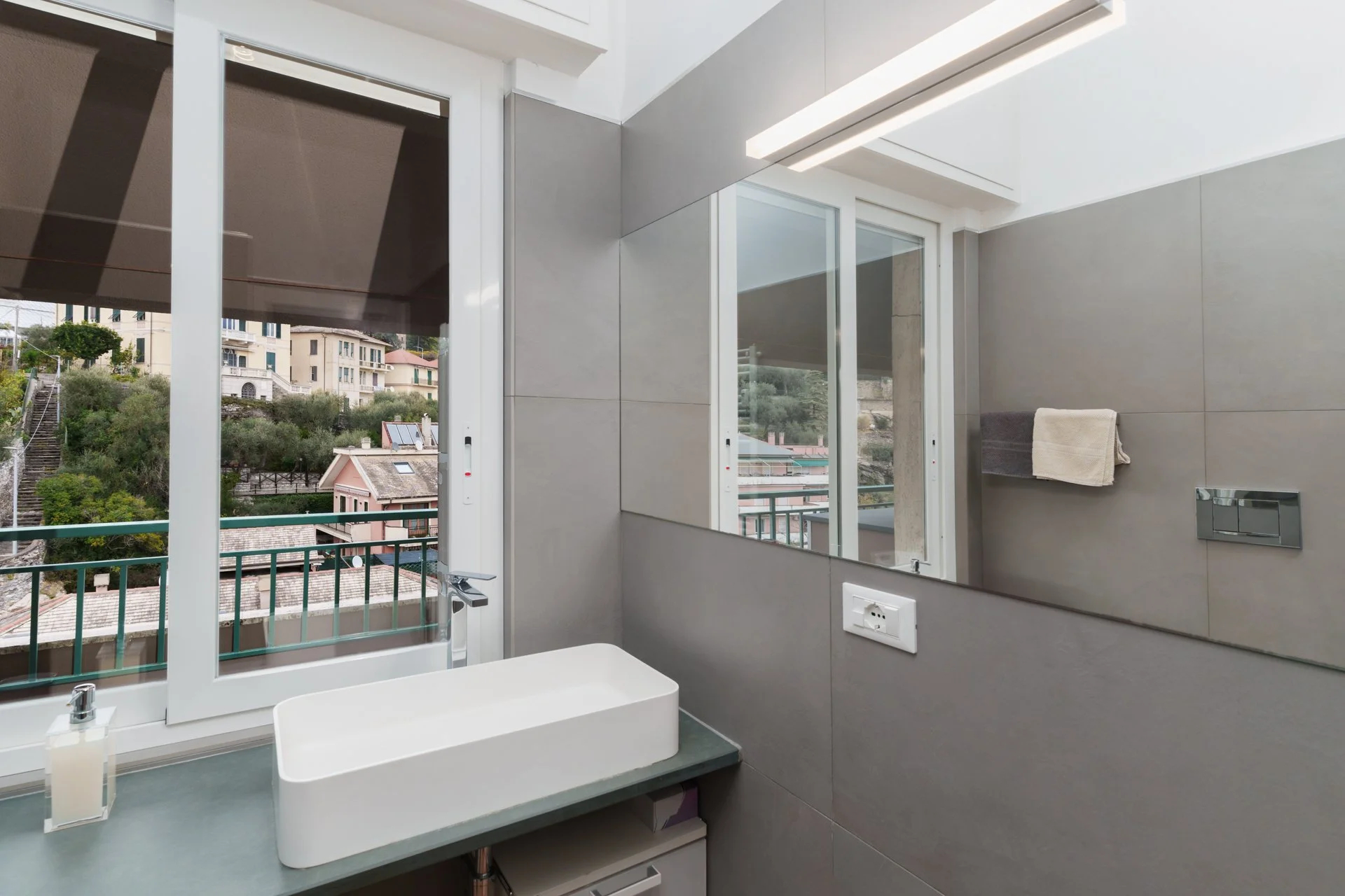 Bagno moderno con finestra che dà su un paesaggio collinare, lavabo rettangolare bianco, specchio e pareti rivestite di piastrelle grigie.