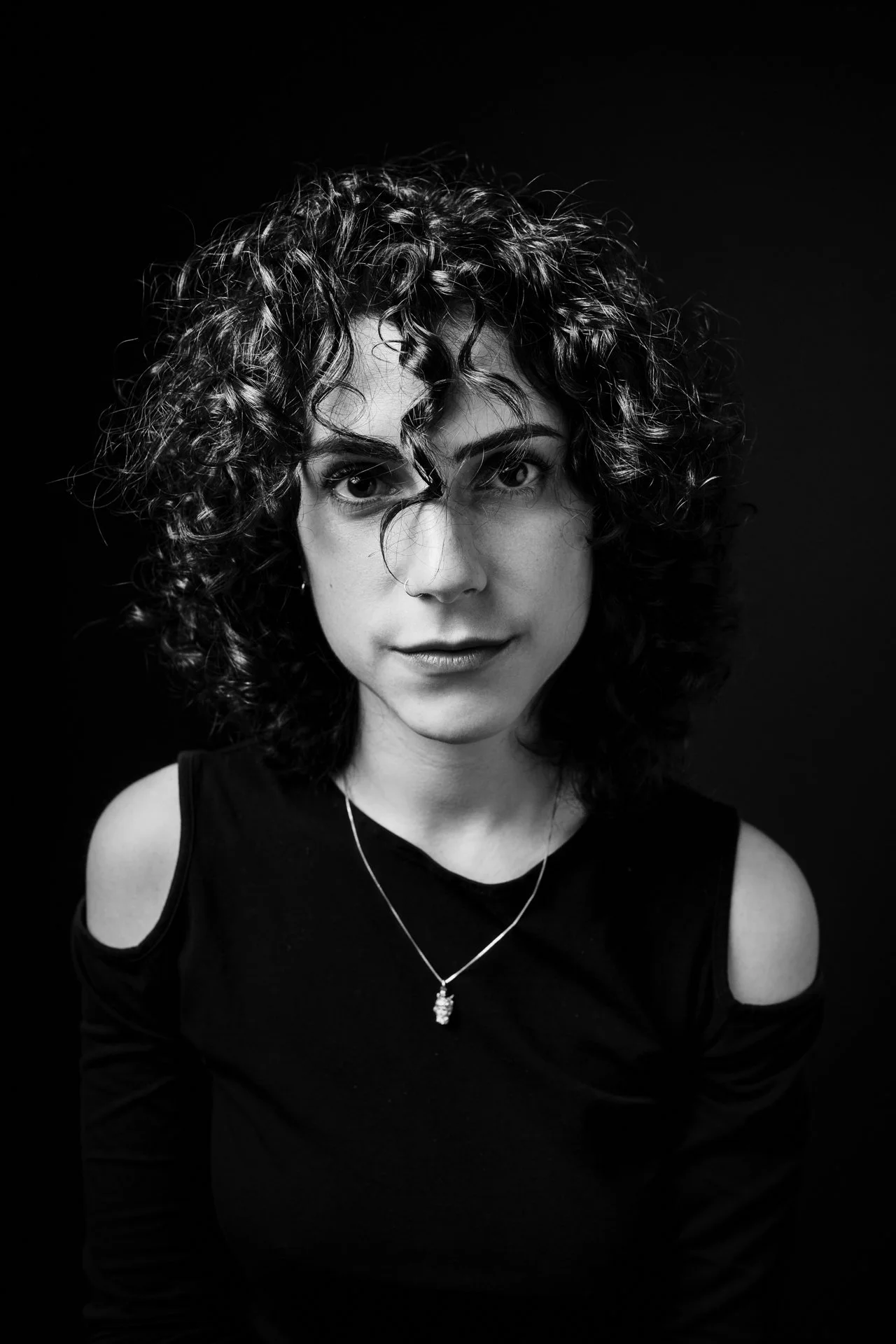 Ritratto in bianco e nero di una donna con capelli ricci, occhi intensi e sorriso sottile, indossa una maglietta con spalline scoperte e una collana.