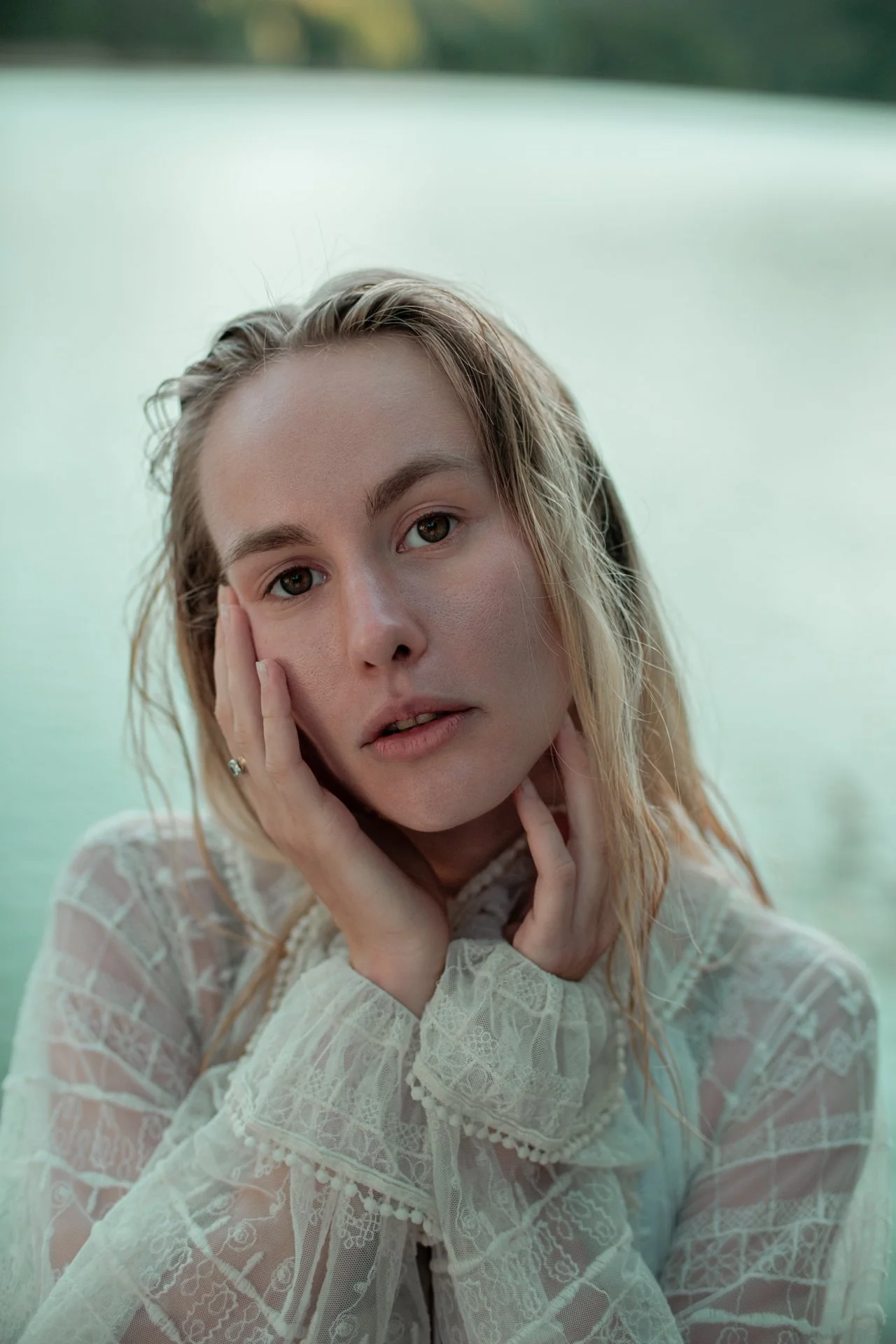 Ragazza con capelli bagnati e vestito bianco con dettagli in pizzo, con una mano sul viso e la testa leggermente inclinata, acqua sullo sfondo.