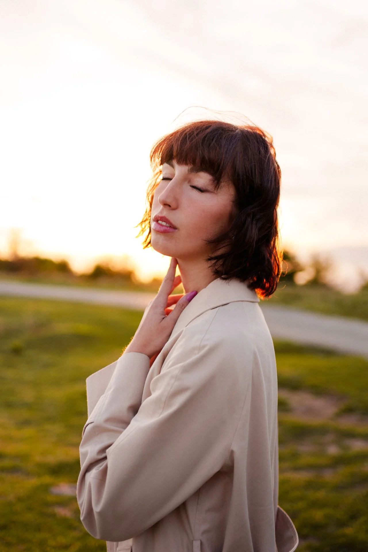 Donna con capelli corti e biondi, occhi chiusi, in abito beige, che si tocca il collo con la mano, sullo sfondo un campo e un tramonto.