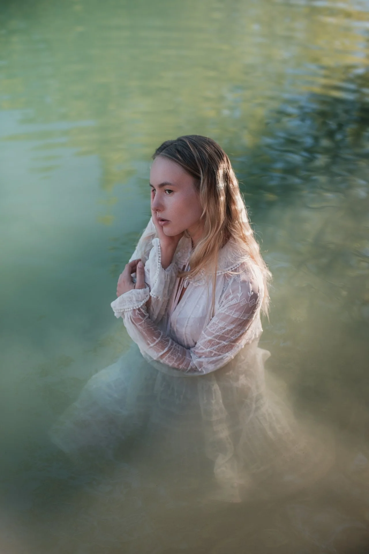 Donna in acqua, con capelli lunghi e abito bianco, che si trova in un ambiente naturale circondato dall'acqua e alberi.