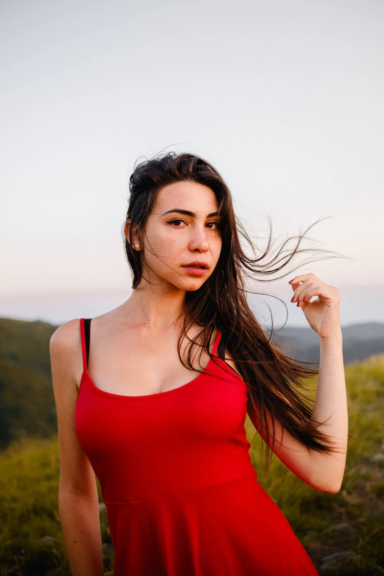 Ragazza con capelli castani e occhi castani, indossa un vestito rosso con spaghette, sfondo naturale con colline e cielo al tramonto.