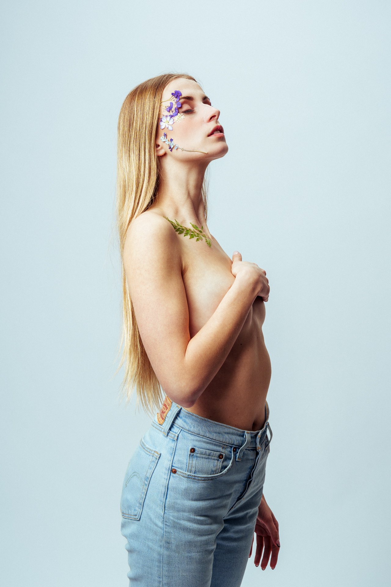 Donna con fiori sulla pelle, copre il petto con il braccio, indossa jeans chiari, sfondo bianco.
