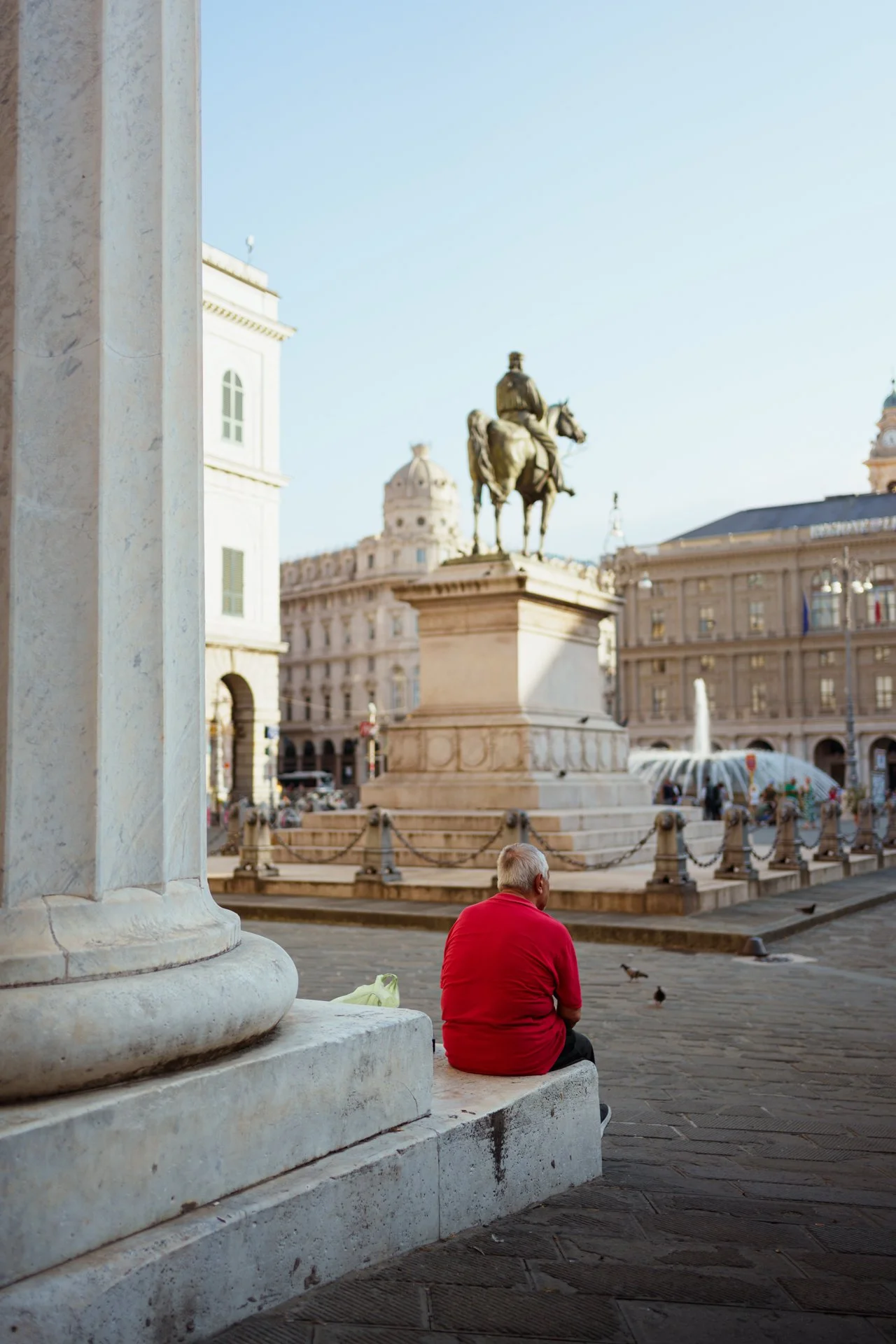 Persona anziana seduta su una panchina di pietra in una piazza con una statua di un uomo a cavallo, fontane e edifici storici sullo sfondo.