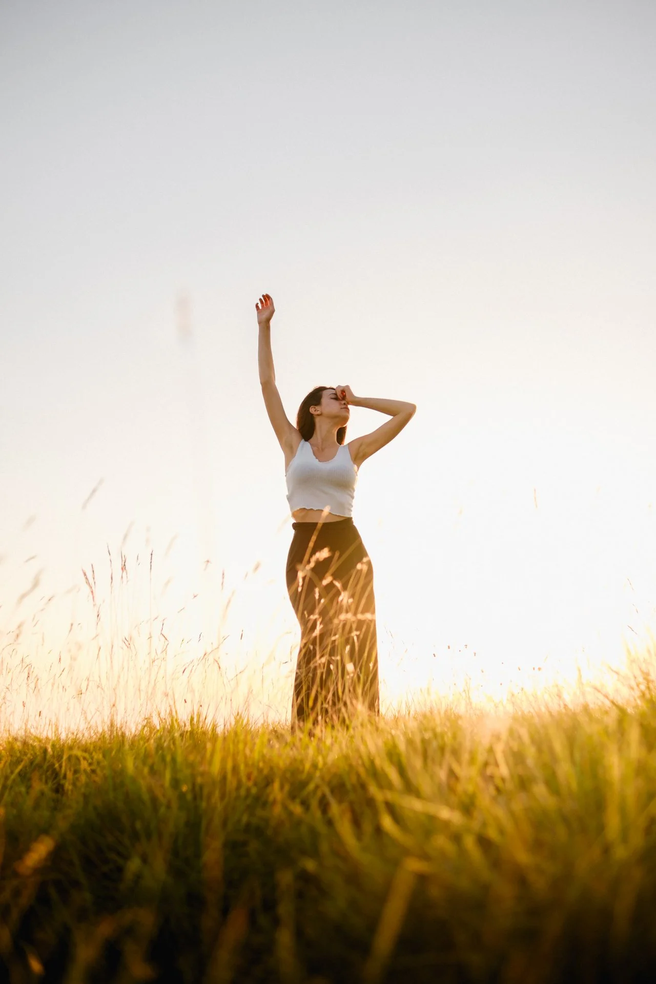 Una donna in piedi in un campo di erba durante il tramonto, con una mano sulla fronte e l'altra alzata verso il cielo.