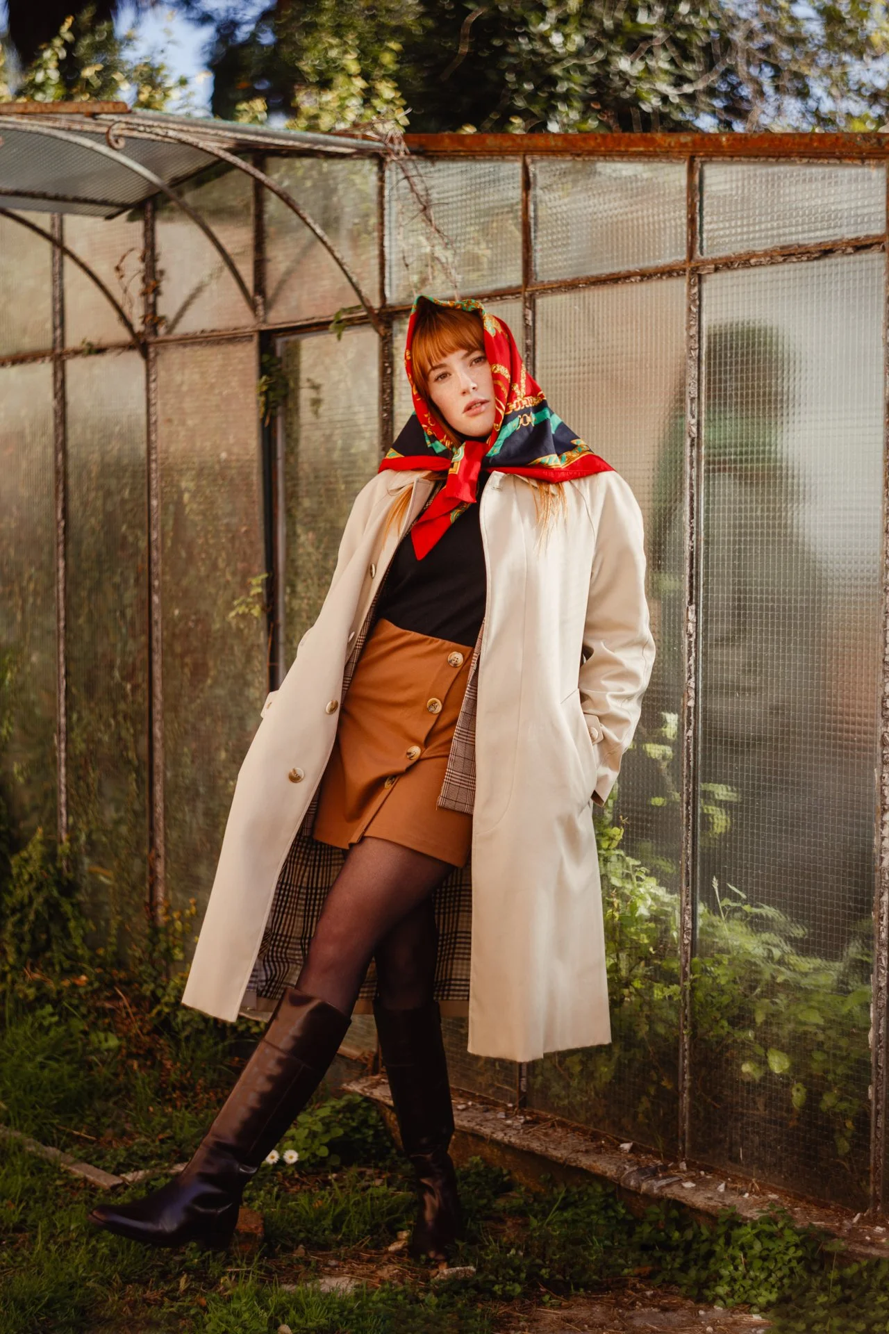 Donna con cappotto beige, gonna cortissima marrone, cappello, stivali neri, e foulard colorato, in un giardino con serra in background.