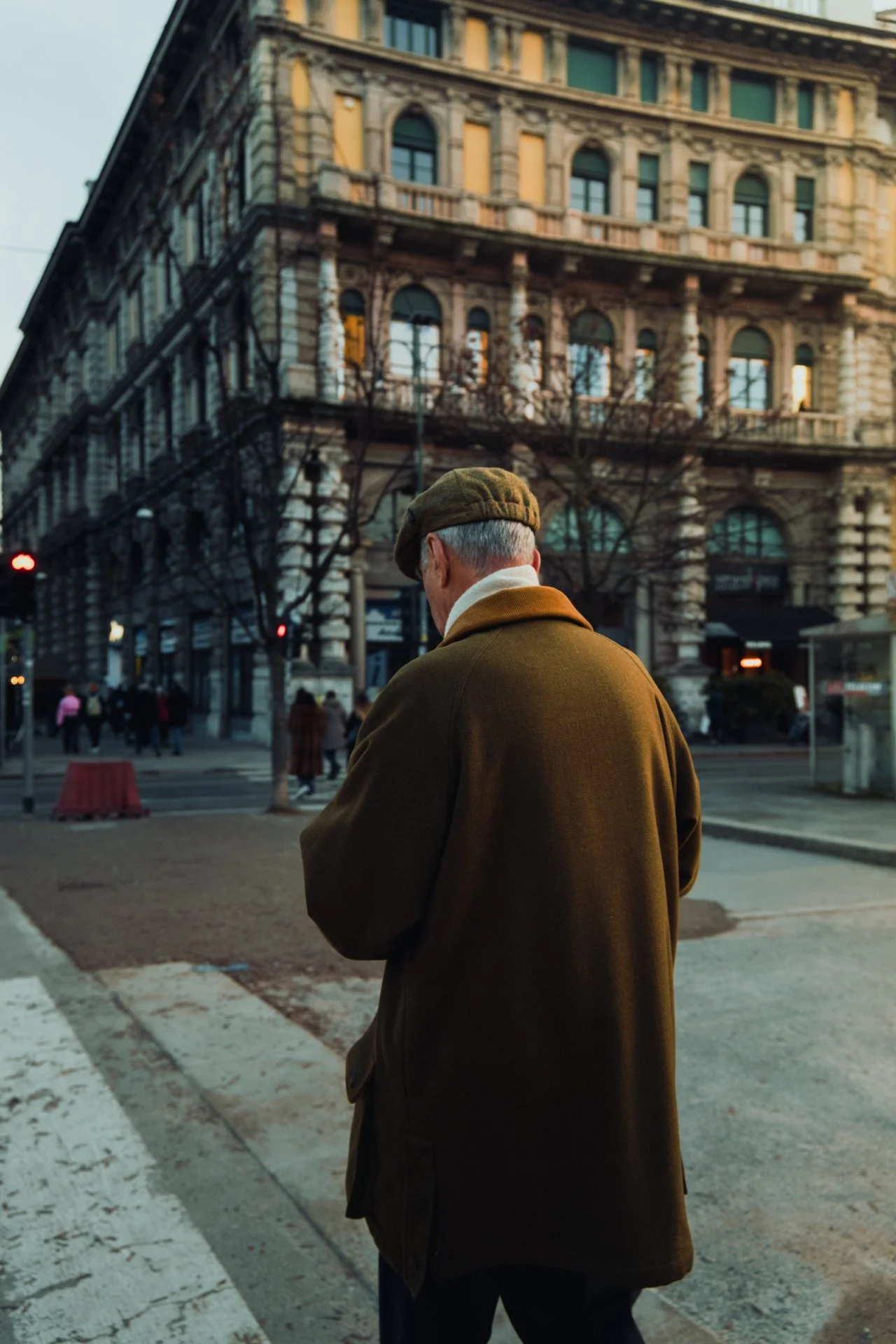 Uomo anziano visto di Schiena, con cappello e cappotto marrone, che attraversa una strada in città, con edifici storici sullo sfondo e alcune persone nella scena.