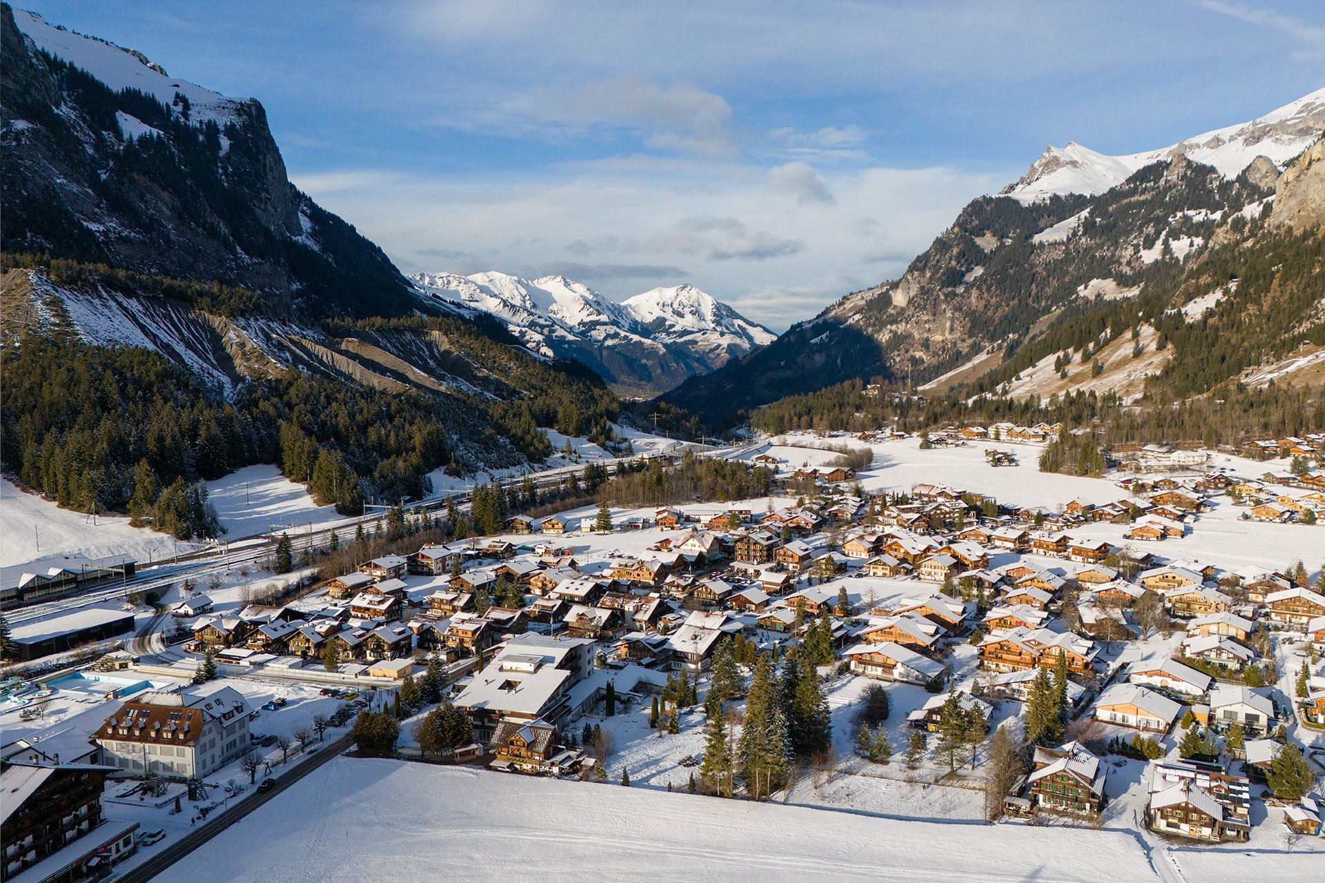 Paesaggio montano invernale con villaggio coperto di neve, case alpine, foreste e montagne innevate sullo sfondo sotto un cielo sereno.