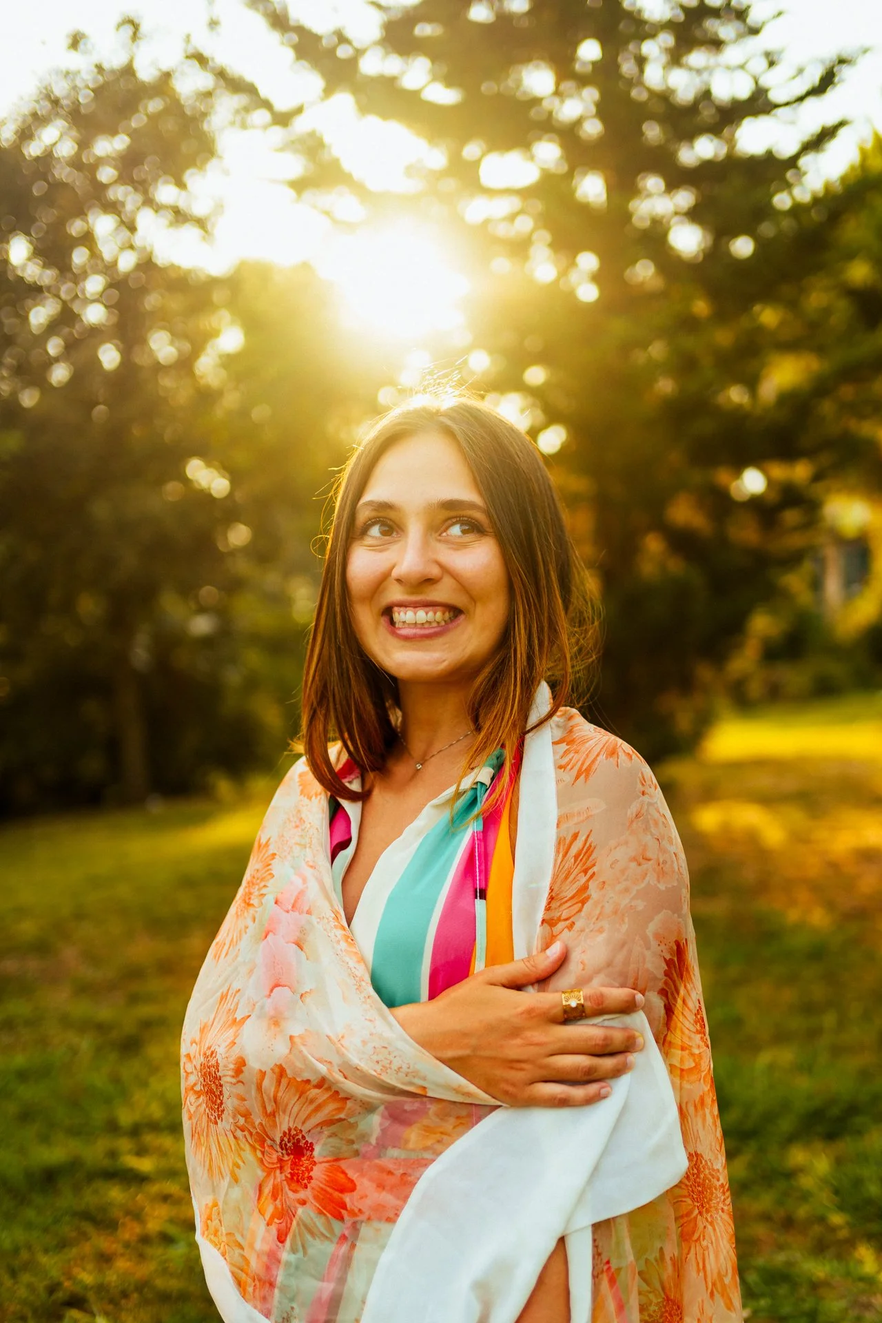 Donna sorridente in un parco al tramonto, con capelli castani e indossa un vestito colorato con un foulard.