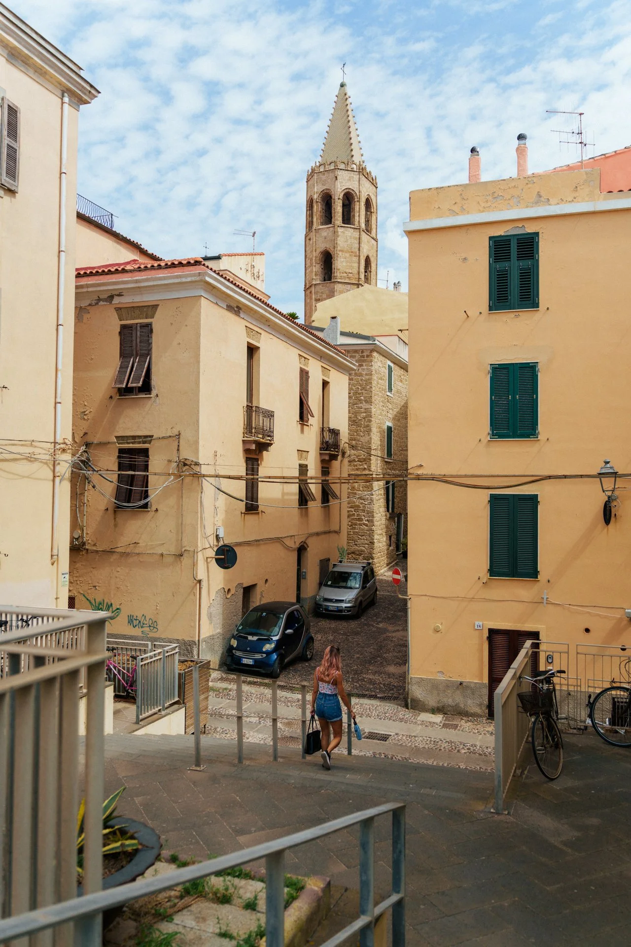 Vista di una strada di un quartiere antico con edifici color beige e gialli, una chiesa con torre campanaria in lontananza, alcune auto parcheggiate, una donna che scende le scale con una borsa e una bici ferma vicino alla ringhiera.