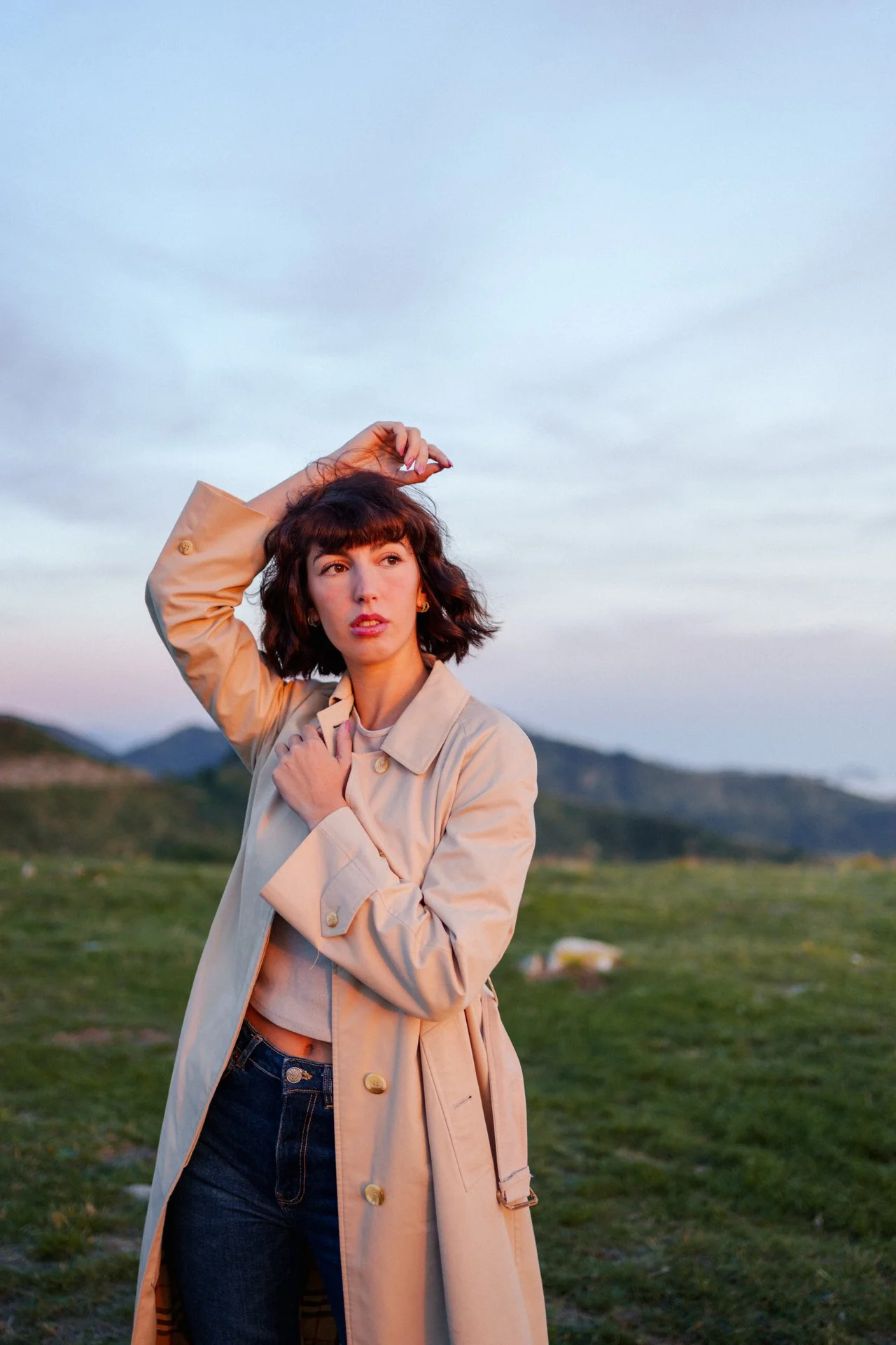Una donna con capelli riccioli corti che indossa un trench beige, in piedi su un paesaggio aperto con colline verdi e un cielo nuvoloso sullo sfondo.