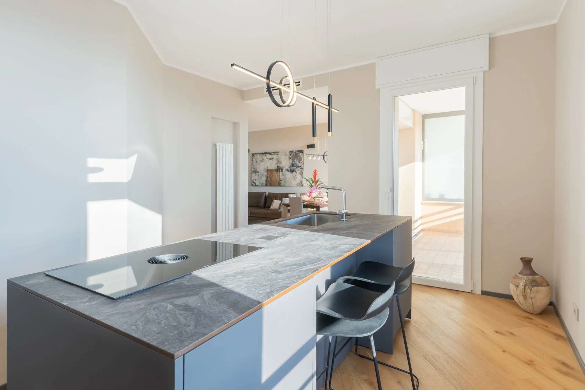 Cucina moderna con isola in marmo grigia e piano cottura a induzione, sedie nere, lampadario artistico sospeso, porta finestra con vista su balcone, pavimento in legno, vaso decorativo in ceramica nell'angolo.