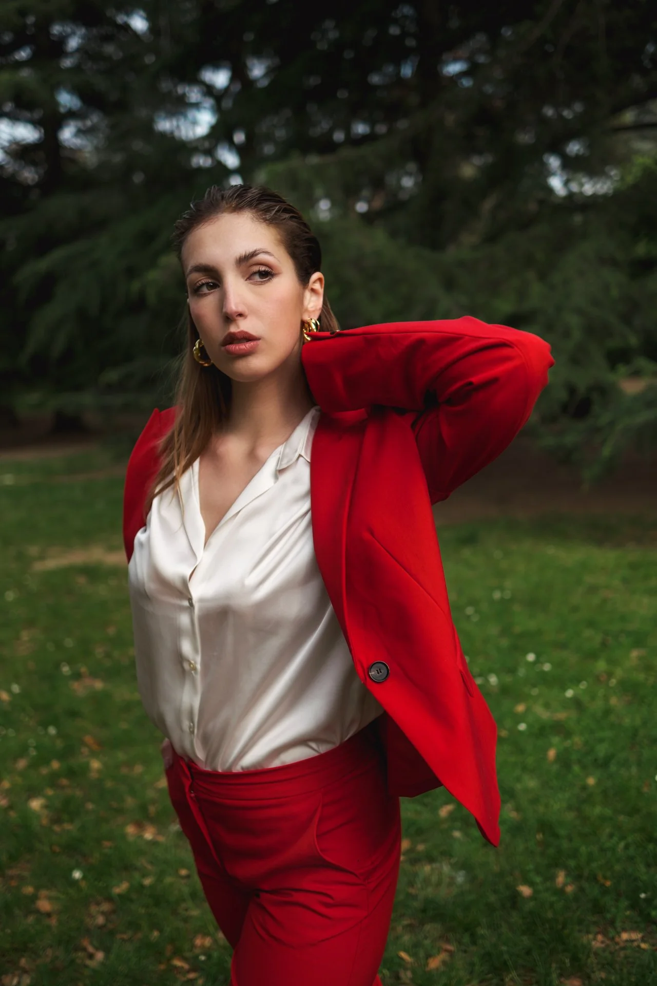 Una donna elegante con un completo rosso e una blusa bianca si trova in un parco circondata da alberi verdi, con un'espressione pensierosa.
