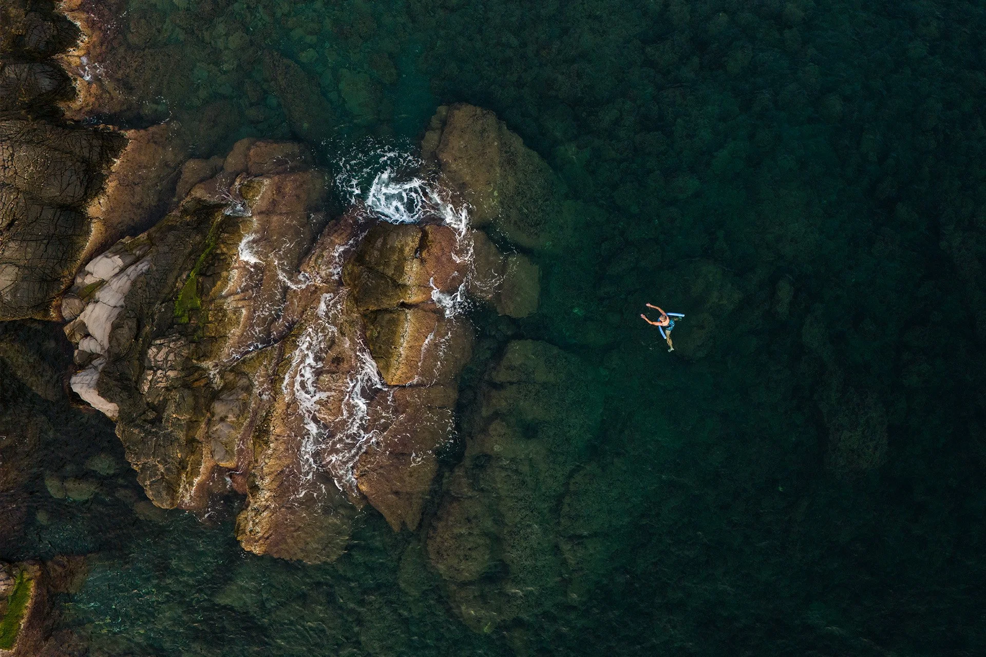 Immagine aerea di una persona che nuota in acque cristalline vicino a rocce e vegetazione lungo la costa.