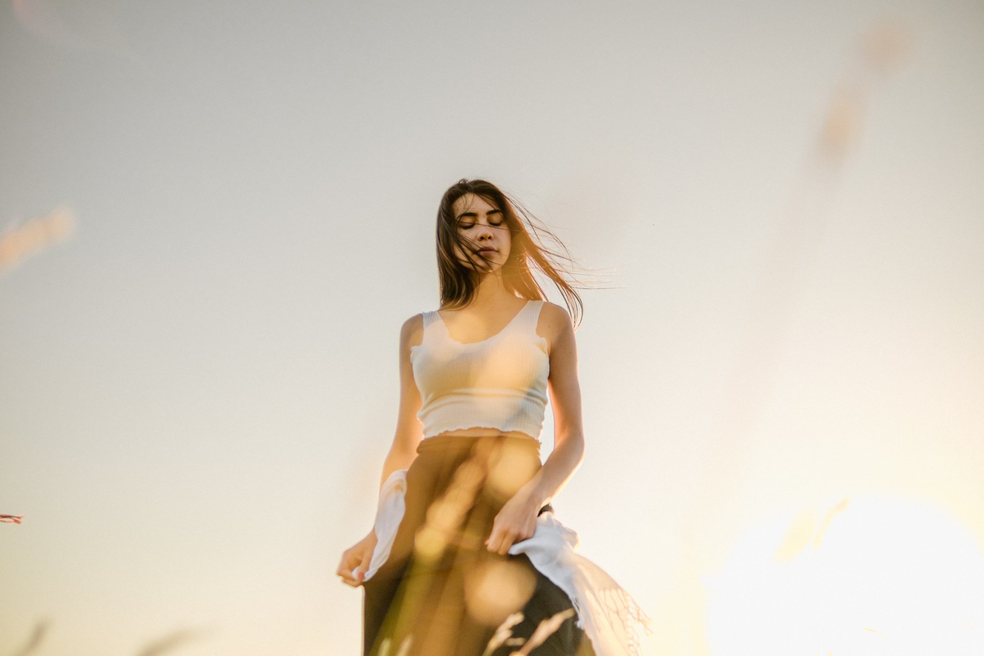 Ragazza che si trova in un campo all'aperto al tramonto, con i capelli mossi dal vento, indossa un top bianco e un vestito nero. La luce calda del tramonto le illumina il viso.
