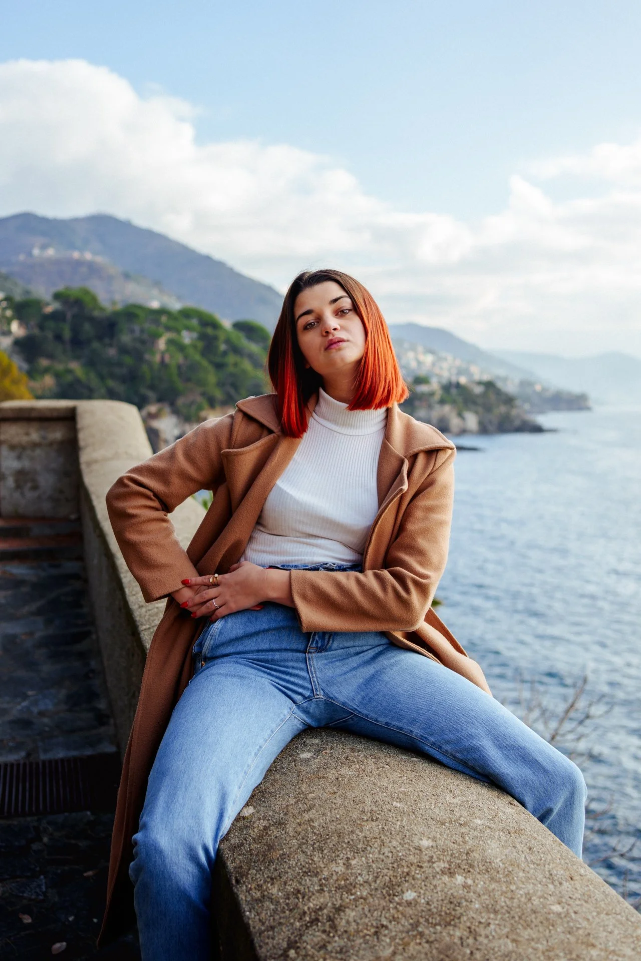 Una giovane donna seduta sulla banchina vicino al mare con colline sullo sfondo, indossa una giacca di mentre e jeans blu, con i capelli rossi.