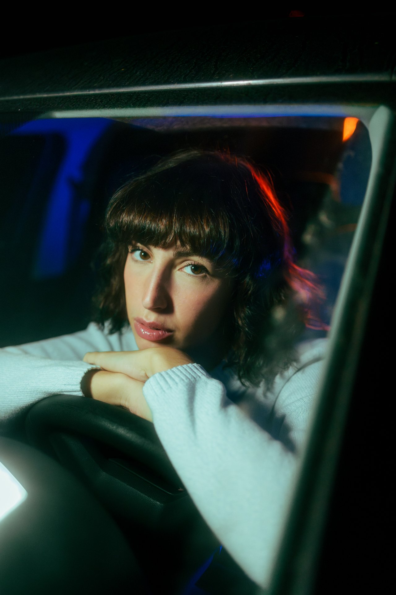 Ragazza con capelli mossi e frangetta, che si appoggia sul volante di un'auto, vista dal parabrezza durante la notte.