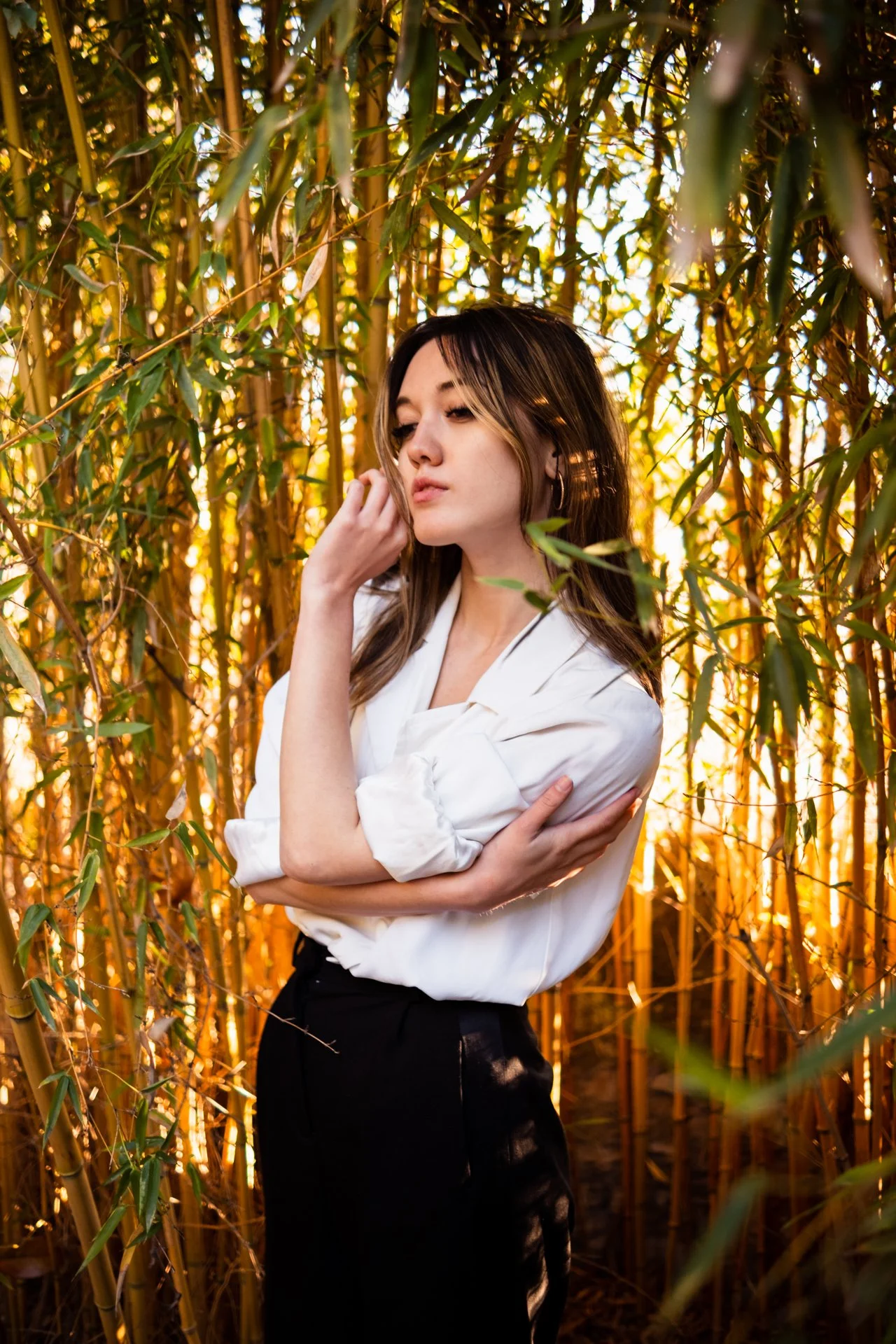Ragazza in piedi tra un campo di canne di bambù, vestita con una camicia bianca e pantaloni neri, con uno sguardo pensieroso, durante il tramonto.
