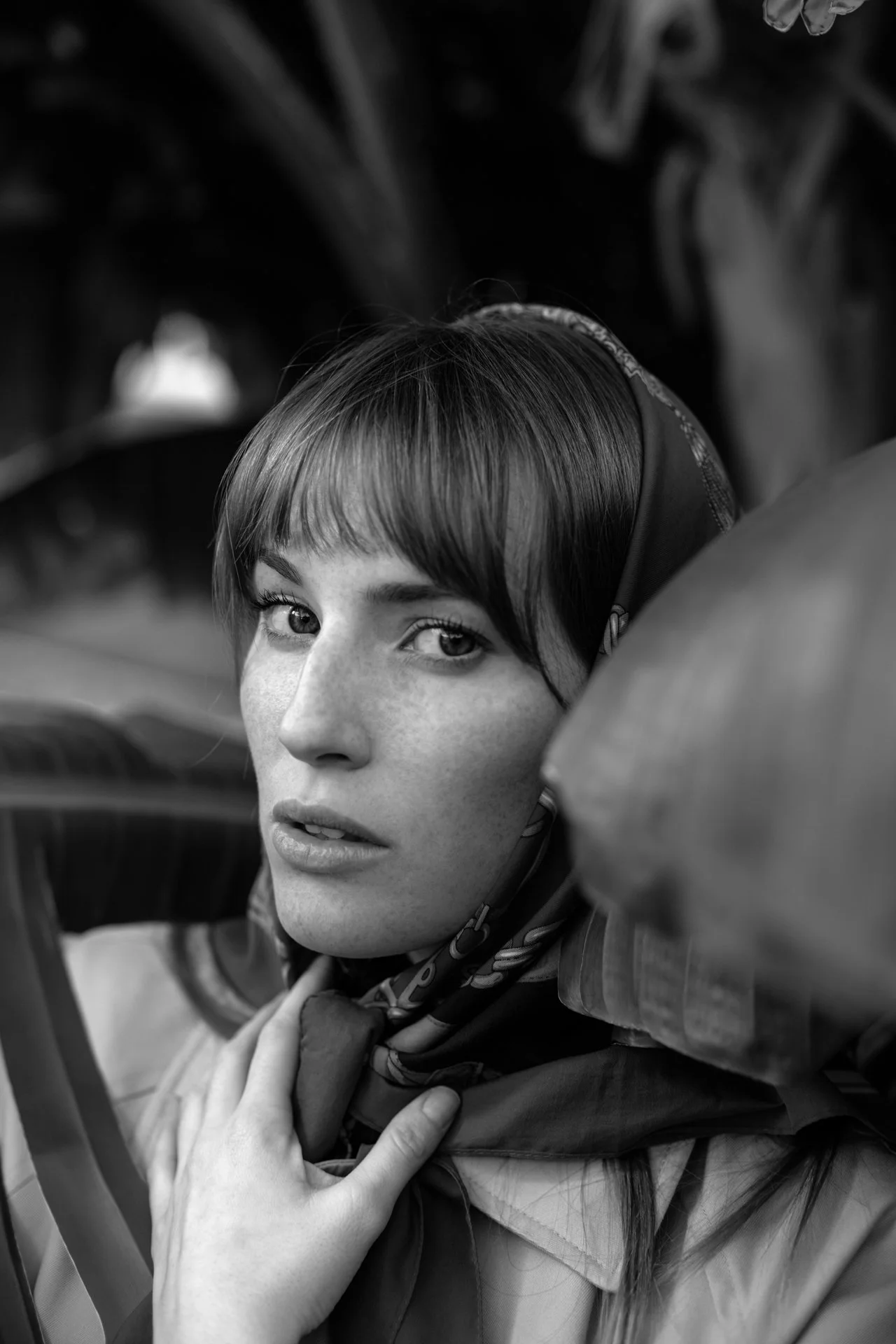 Ragazza con capelli corti e occhi chiari, vestita con una sciarpa, in uno scatto in bianco e nero con sfondo naturale.