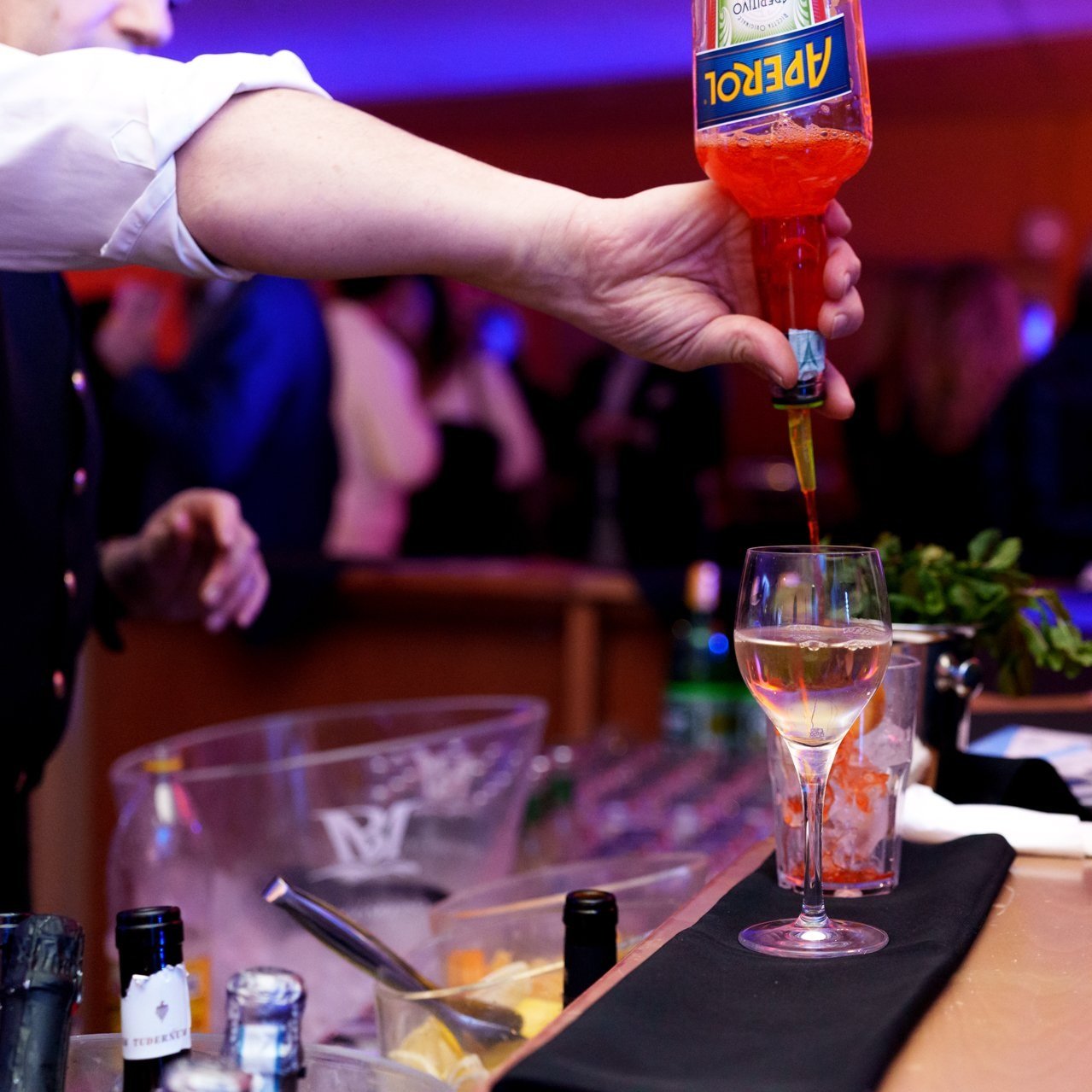 Una mano sta versando un cocktail rosa da una bottiglia di Aperol in un bicchiere di vino, con altre bottiglie e utensili sul bancone da bar.