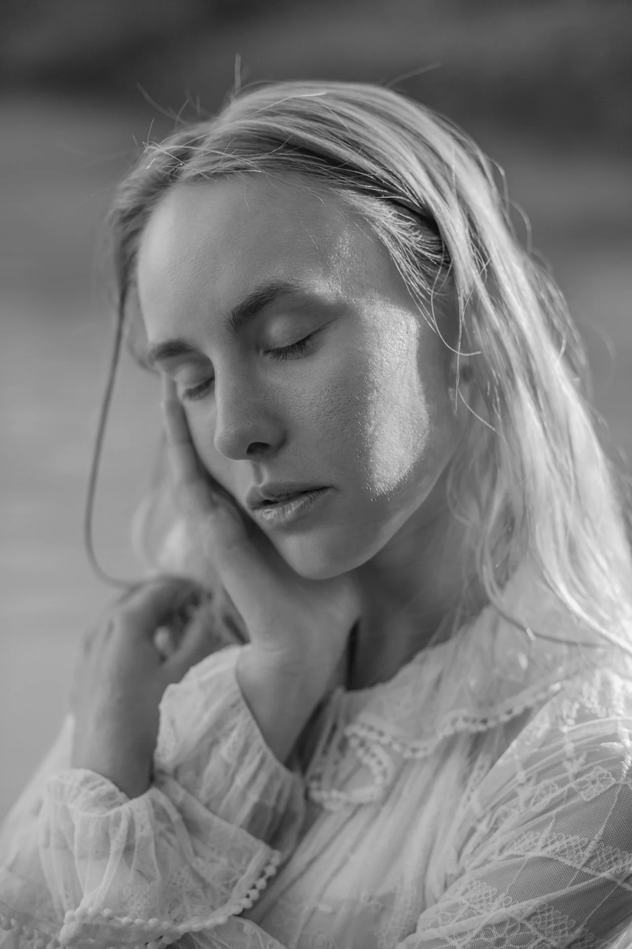 Ritratto in bianco e nero di una donna con i capelli biondi, occhi chiusi, che tiene una mano al lato del collo, con un vestito di pizzo con dettagli.