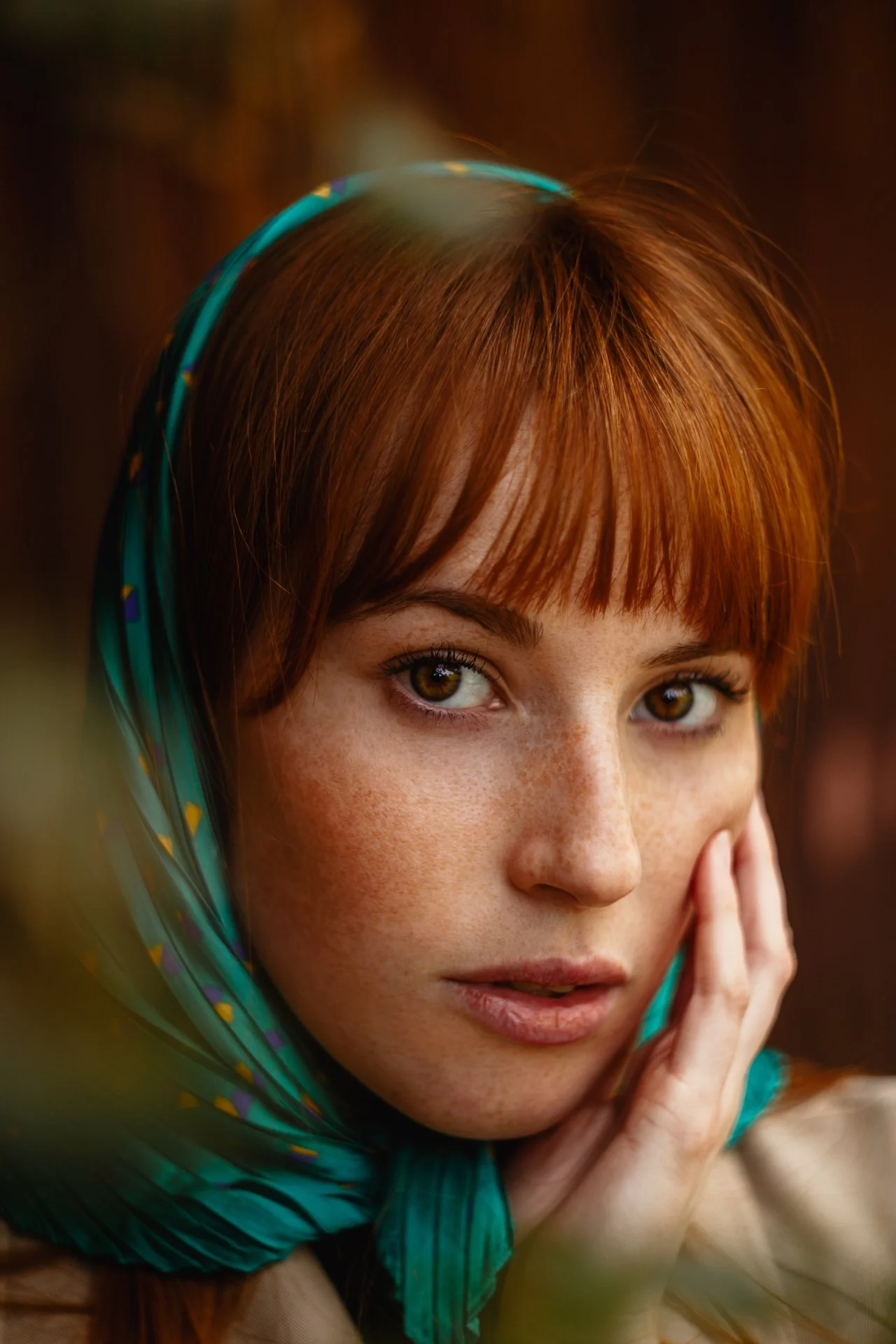 Primissima piano di donna con capelli rossi, occhi castani, e pelle chiara, che indossa una sciarpa verde con disegni colorati, toccandosi il viso con una mano, sfondo sfocato.