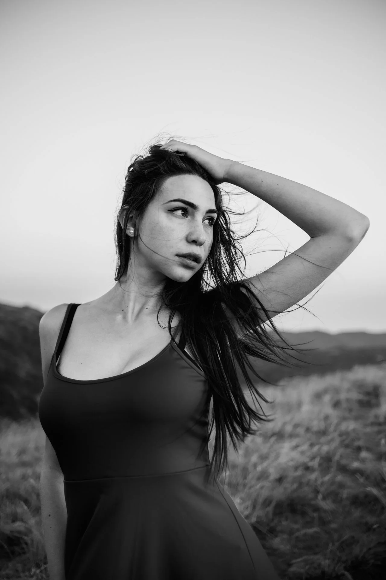 Ragazza con lunghi capelli neri, indossa un vestito senza maniche, in un paesaggio naturale con colline, vista in bianco e nero, che guarda verso sinistra con una mano sulla testa.