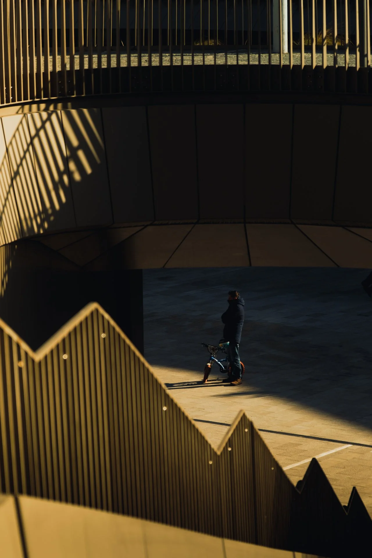 Persona con giacca nera e cappello vicino a una bici, sotto un ponte o una struttura architettonica moderna con ringhiere dorate e ombre nette create dalla luce solare.
