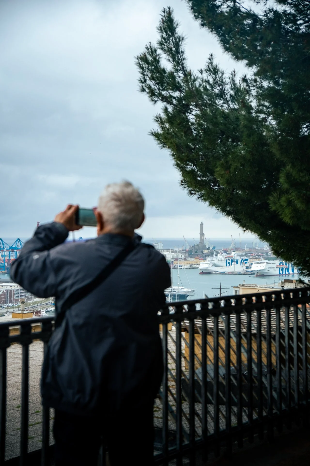 Persona anziana che scatta una fotografia del porto con una torre e navi in lontananza, durante un giorno nuvoloso.