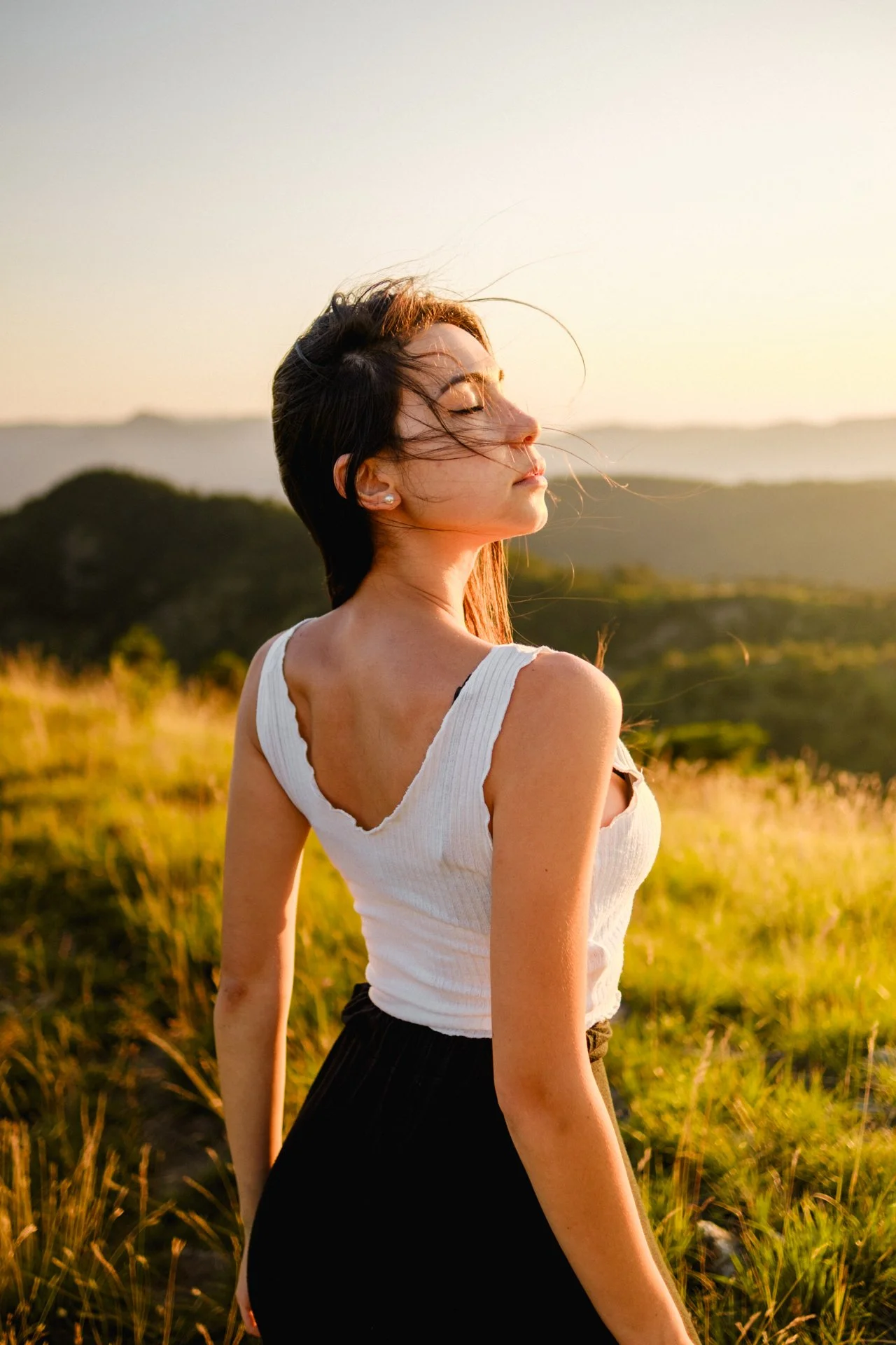 Una giovane donna con i capelli lunghi e scuri, vestita con una maglietta bianca senza maniche e una gonna nera, si trova in un campo verde al tramonto con occhi chiusi e dietro un paesaggio collinare.